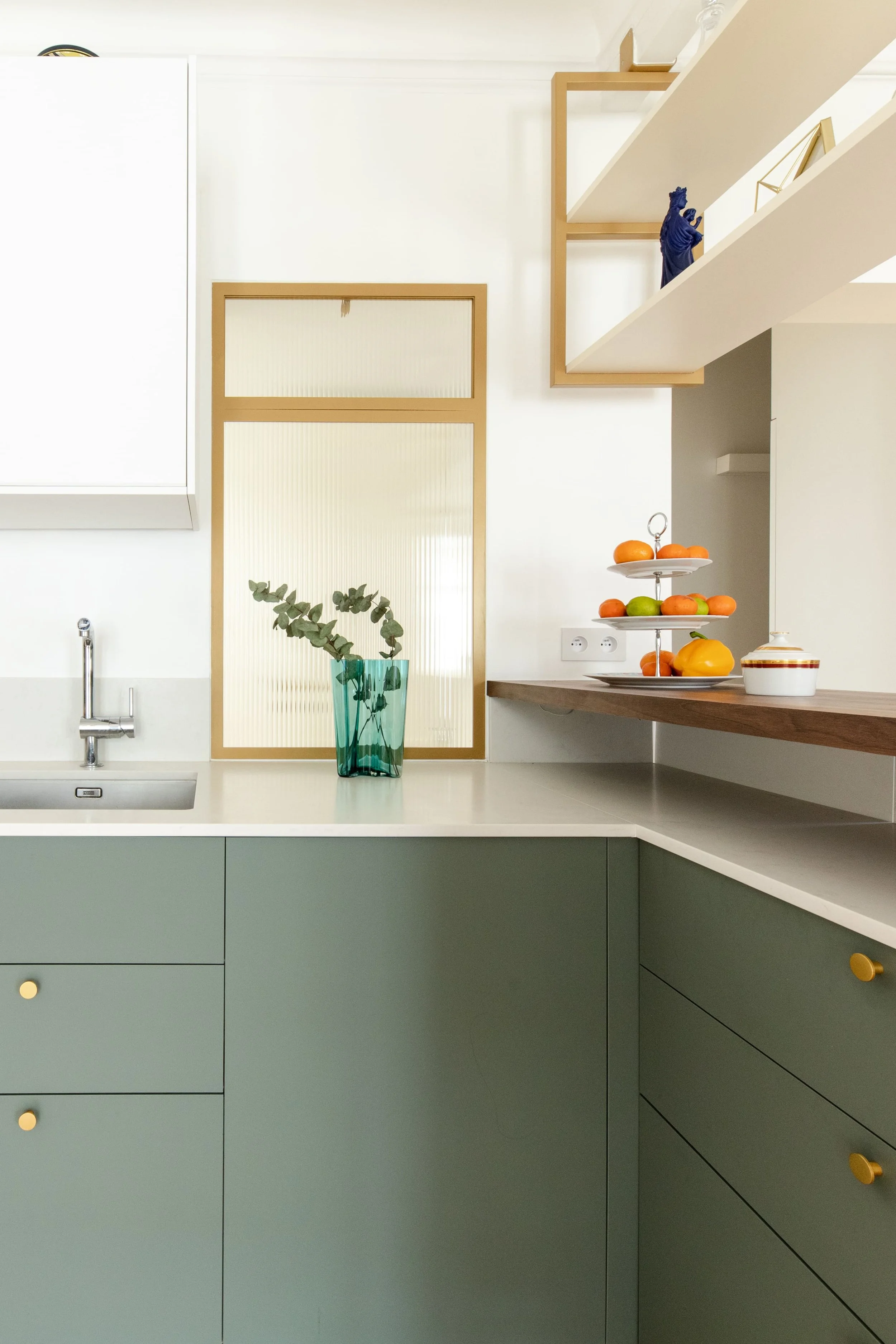 Coin de cuisine avec comptoir, vasque, vase avec feuillage, verrière dorée avec verre fluté,  étagères avec décoration, panier de fruits et boîte