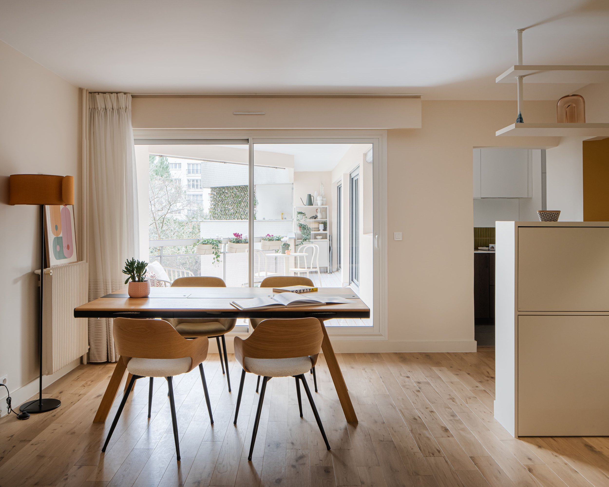 Salon lumineux avec une table en bois entourée de cinq chaises, vue sur un balcon avec des pots de fleurs, décor moderne avec meubles neutres et sol en bois.