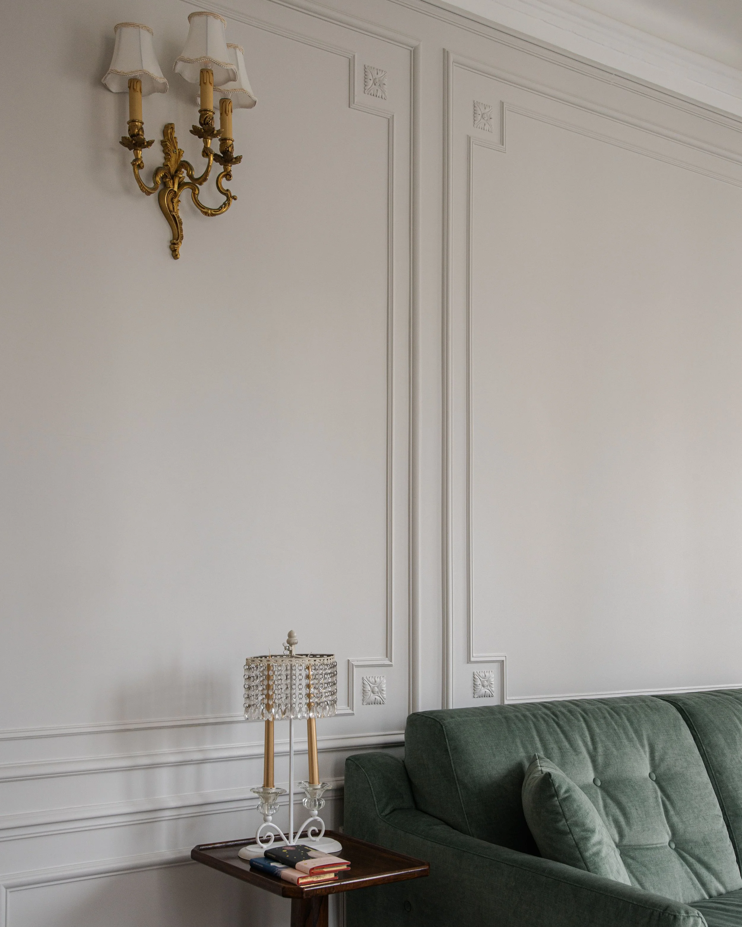 Salon avec mur blanc orné de moulures, lampe de table avec abat-jour en cristaux, canapé vert en velours, et applique murale dorée avec trois bras et abat-jour blanc.