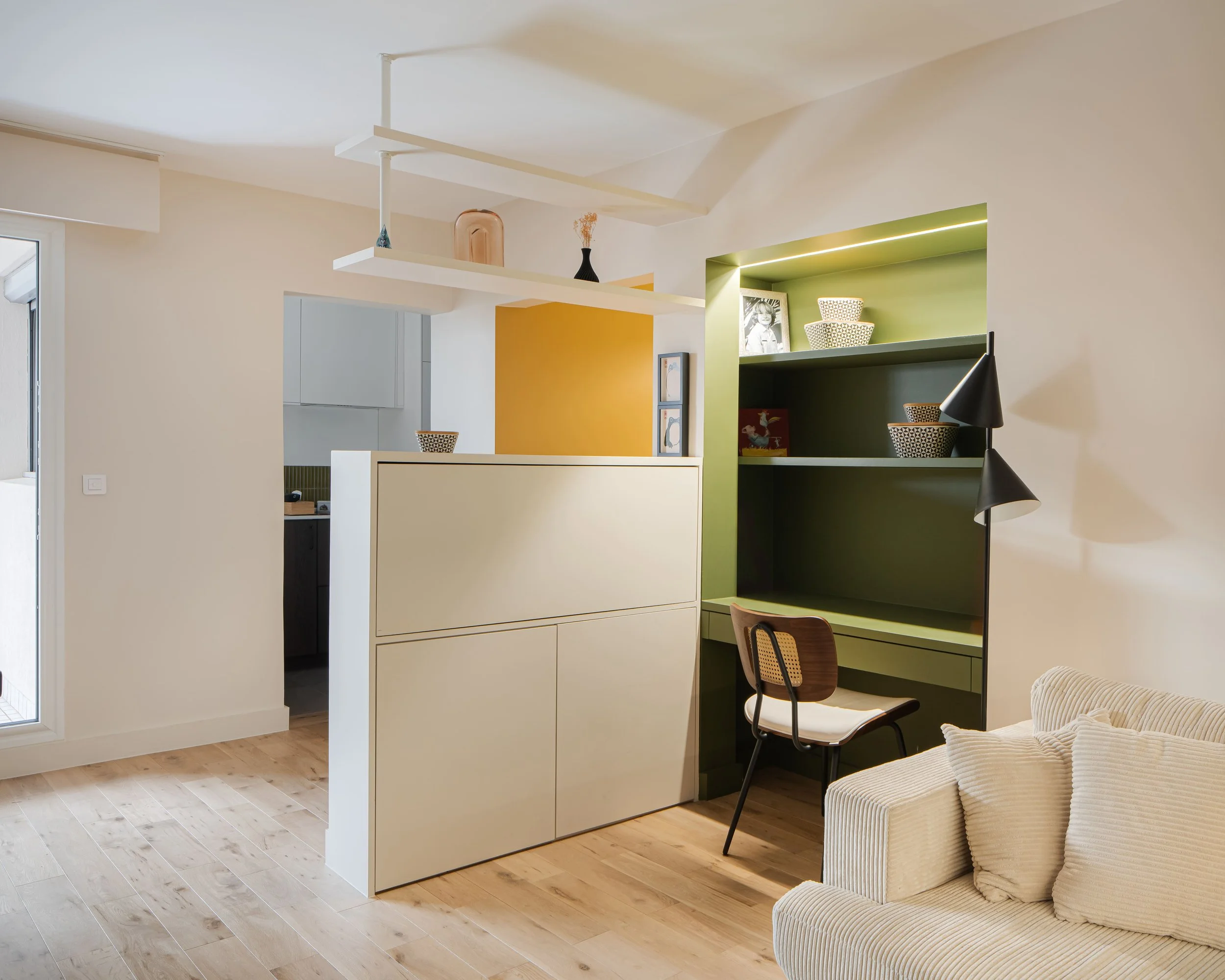 Salon avec une étagère verte, une chaise en bois avec coussin blanc, un canapé beige à coussins, un lampadaire noir à deux têtes, et une cloison blanche séparant partiellement la cuisine.