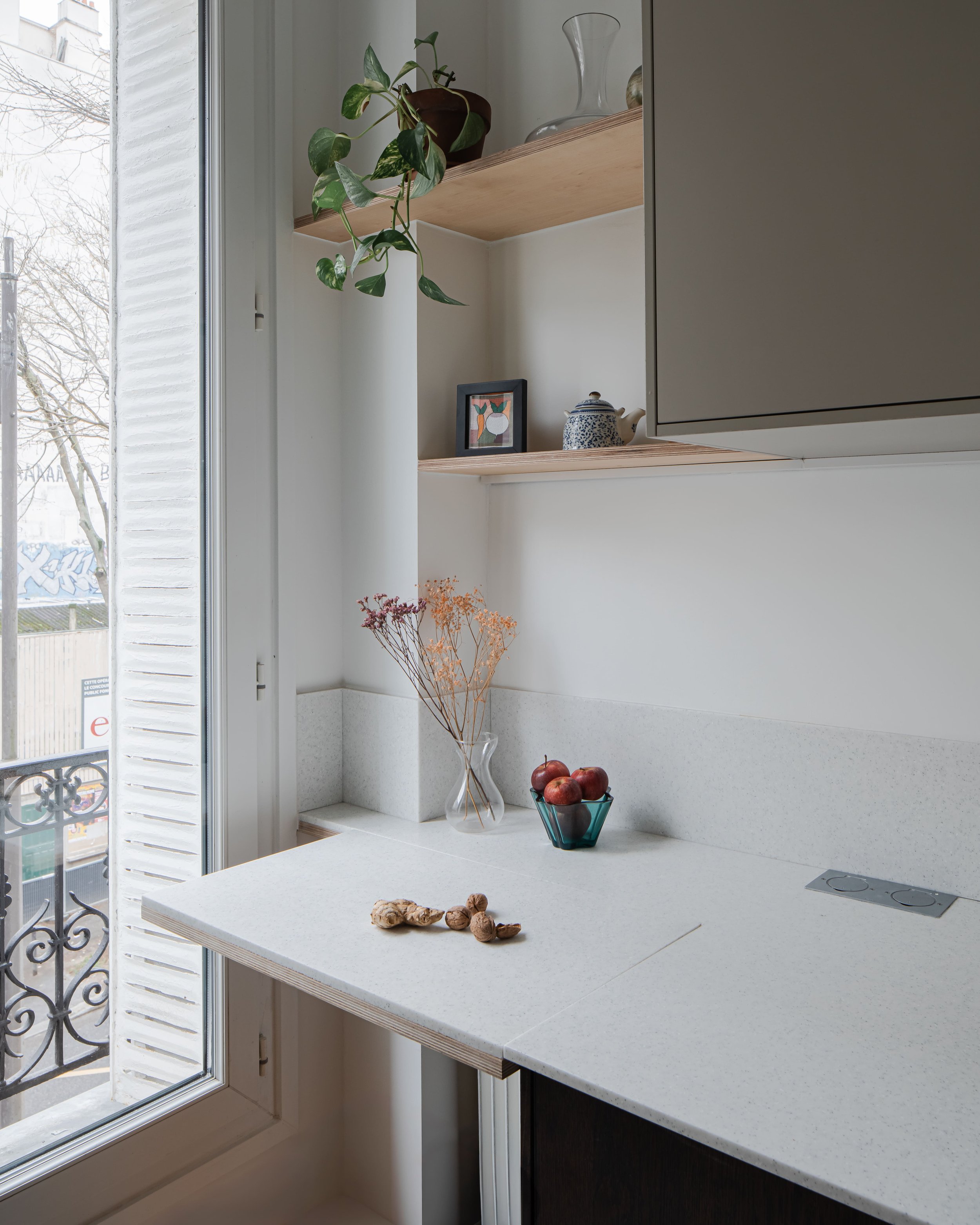 Une cuisine moderne avec un comptoir blanc, quelques noix, une coupe d'herbes, et une fenêtre laissant entrer la lumière naturelle.