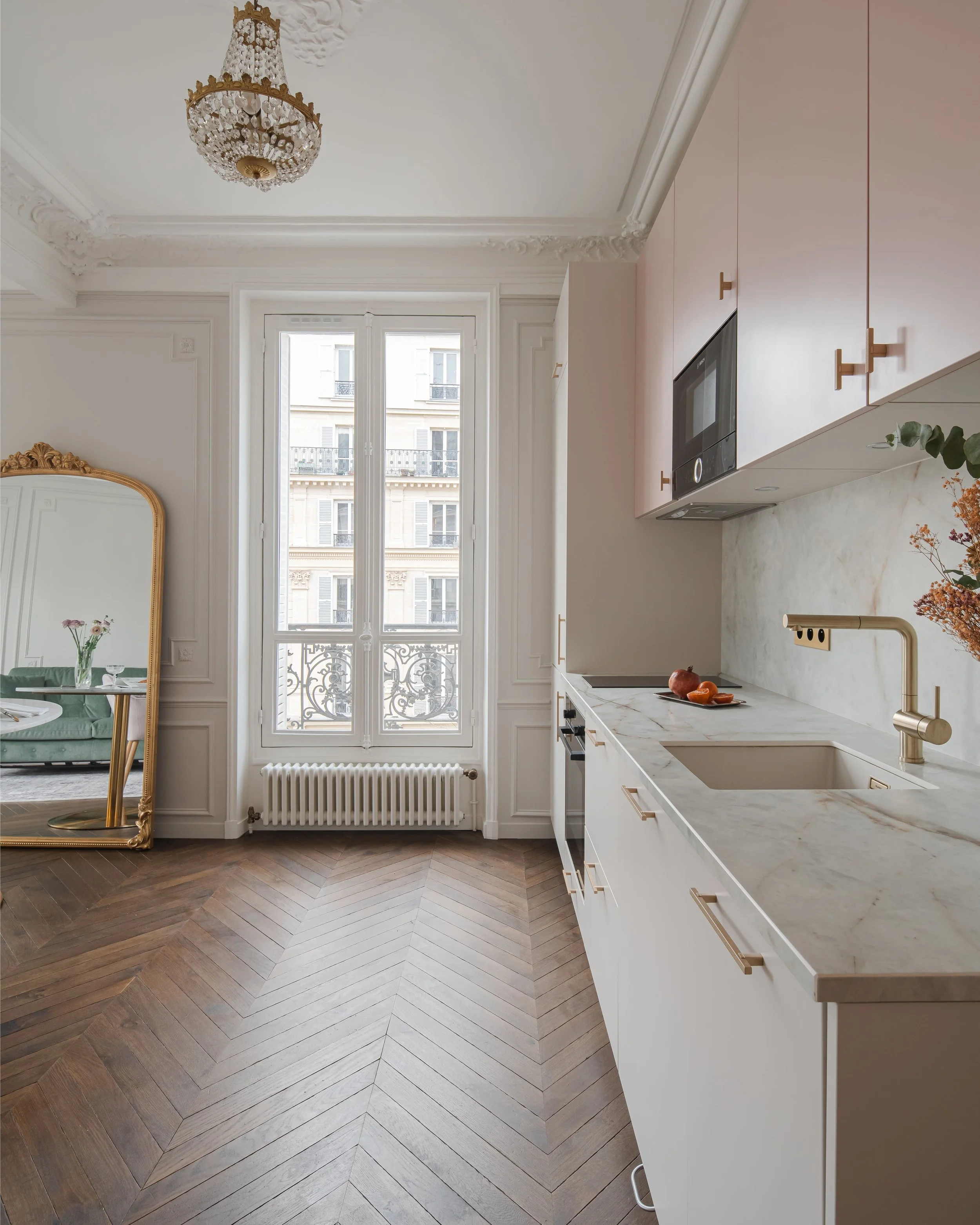 Cuisine moderne avec plan de travail en marbre, armoires pastel, et grande fenêtre avec vue sur un immeuble haussmannien, parquet en chevrons, porte-miroir doré, et lustre chandelier.