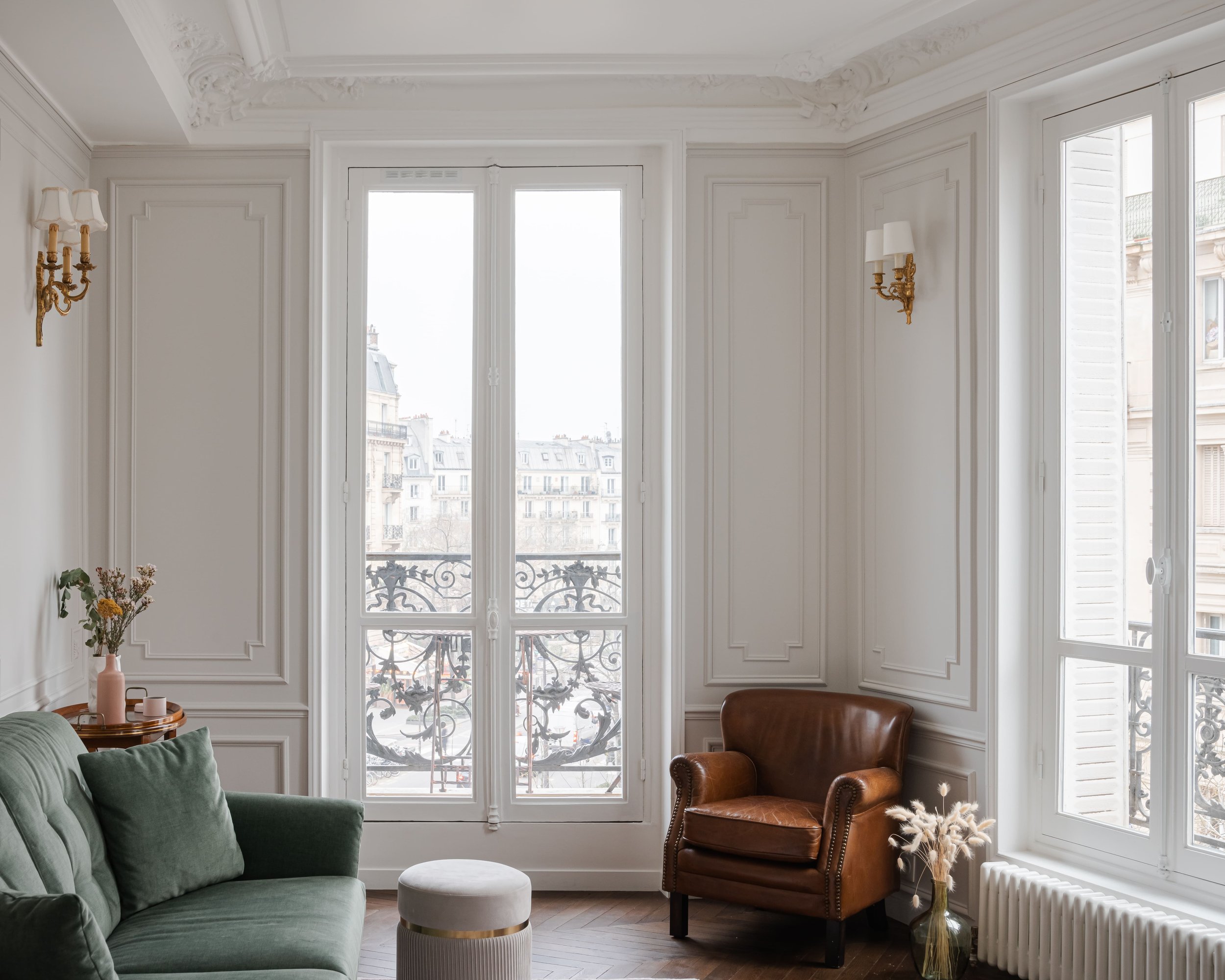 Salon élégant avec grandes fenêtres, canapé vert, fauteuil en cuir marron, décor classique, bouquet de fleurs, et vue urbaine.