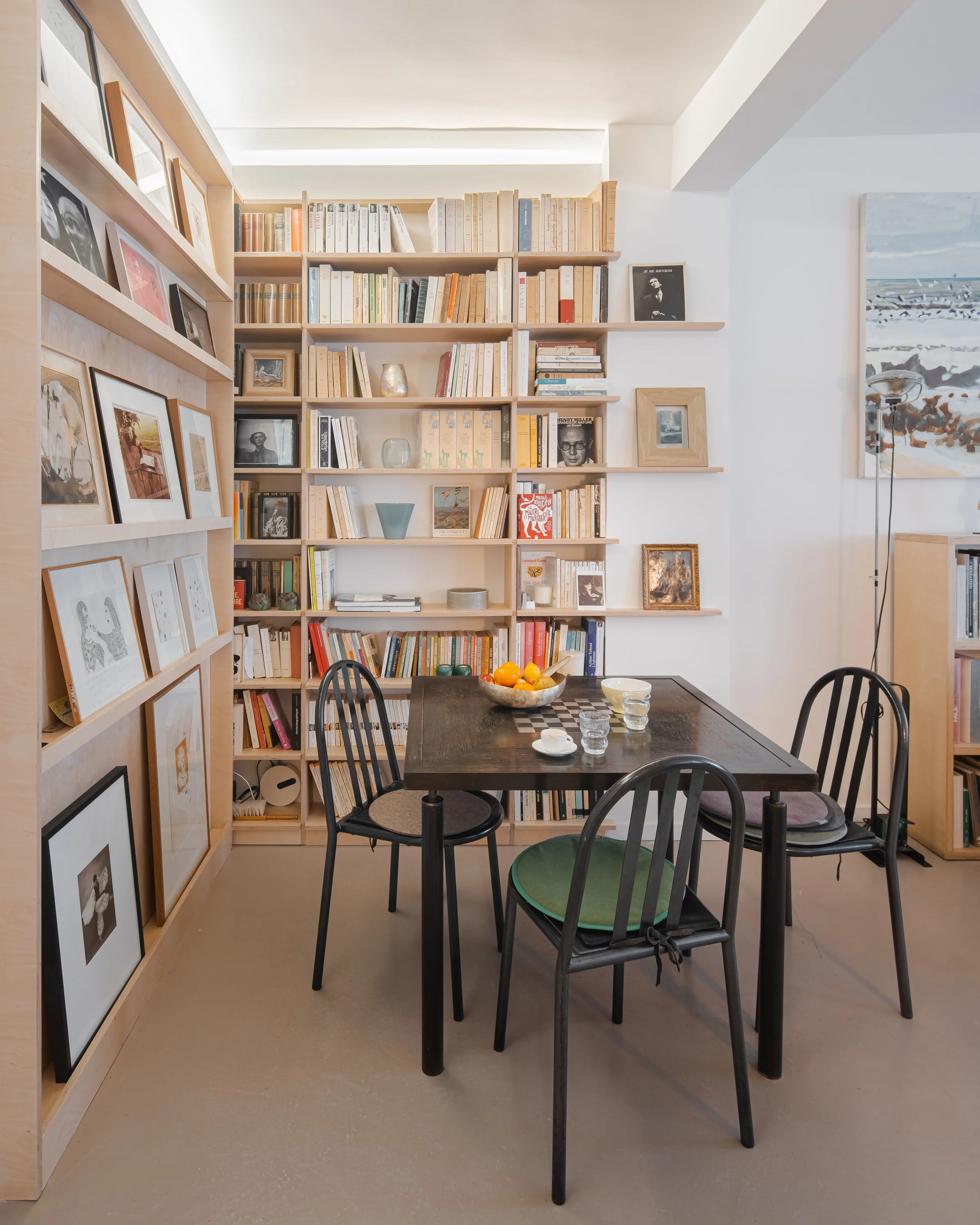 Salle à manger avec un mur de bibliothèque rempli de livres, une table en bois noire avec des chaises noires, des verres et un bol de fruits, lampe sur le côté, tableau de paysage sur le mur à droite.