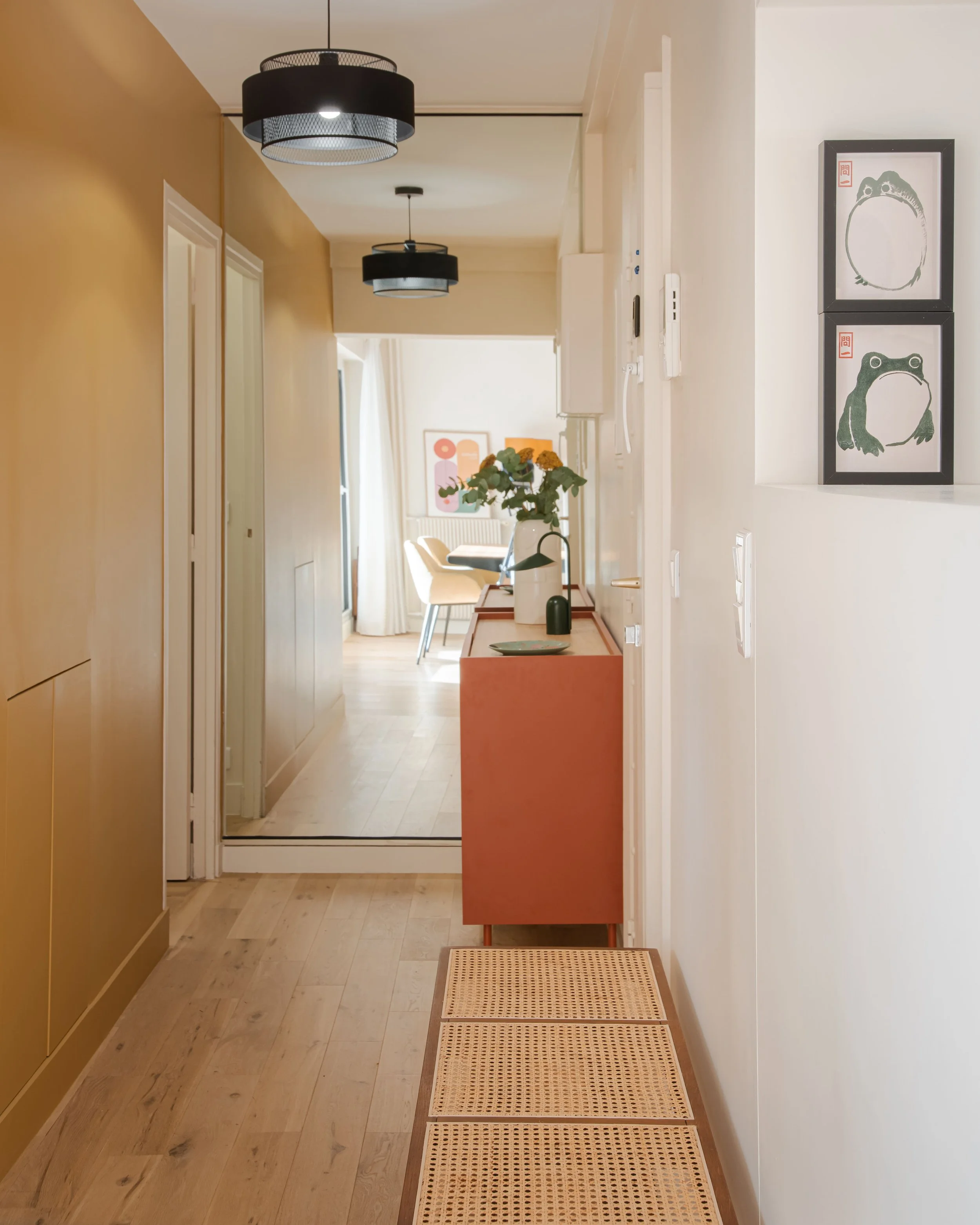 Intérieur lumineux d'un appartement avec un couloir décoré de tableaux de grenouilles, une console orange avec des vases et des fleurs, et une salle à manger au bout du couloir.