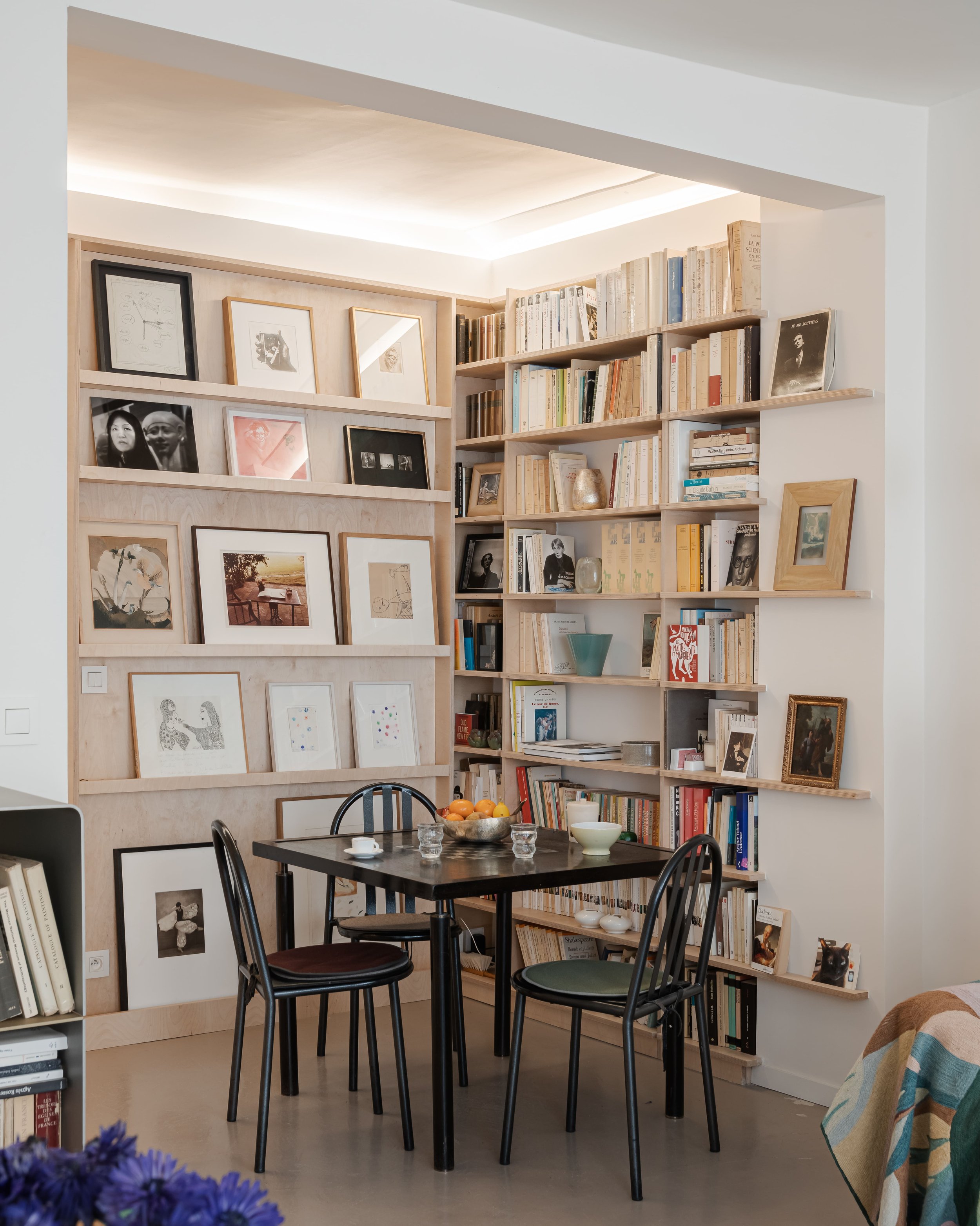 Une bibliothèque intérieure avec étagères en bois remplies de livres, de cadres et d'objets décoratifs. Un tapis blanc, une table noire avec quatre chaises autour, et des fruits dans un bol en métal sur la table.