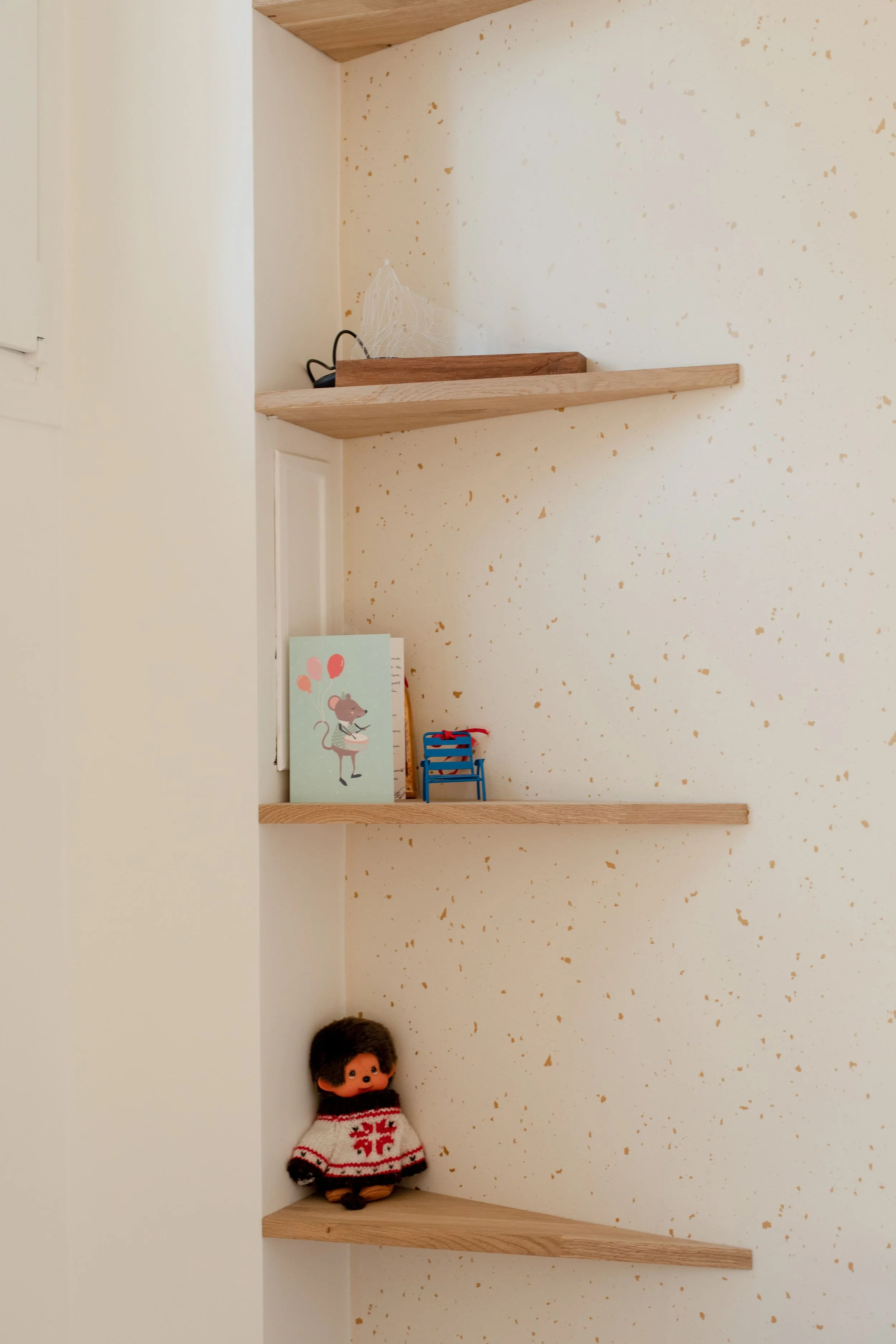 Étagères en bois fixées sur un mur clair avec des objets pour enfant, dont un carnet avec un rat dessiné, une petite chaise bleue, une poupée en laine avec un uniforme blanc et rouge, et un décor de Noël.