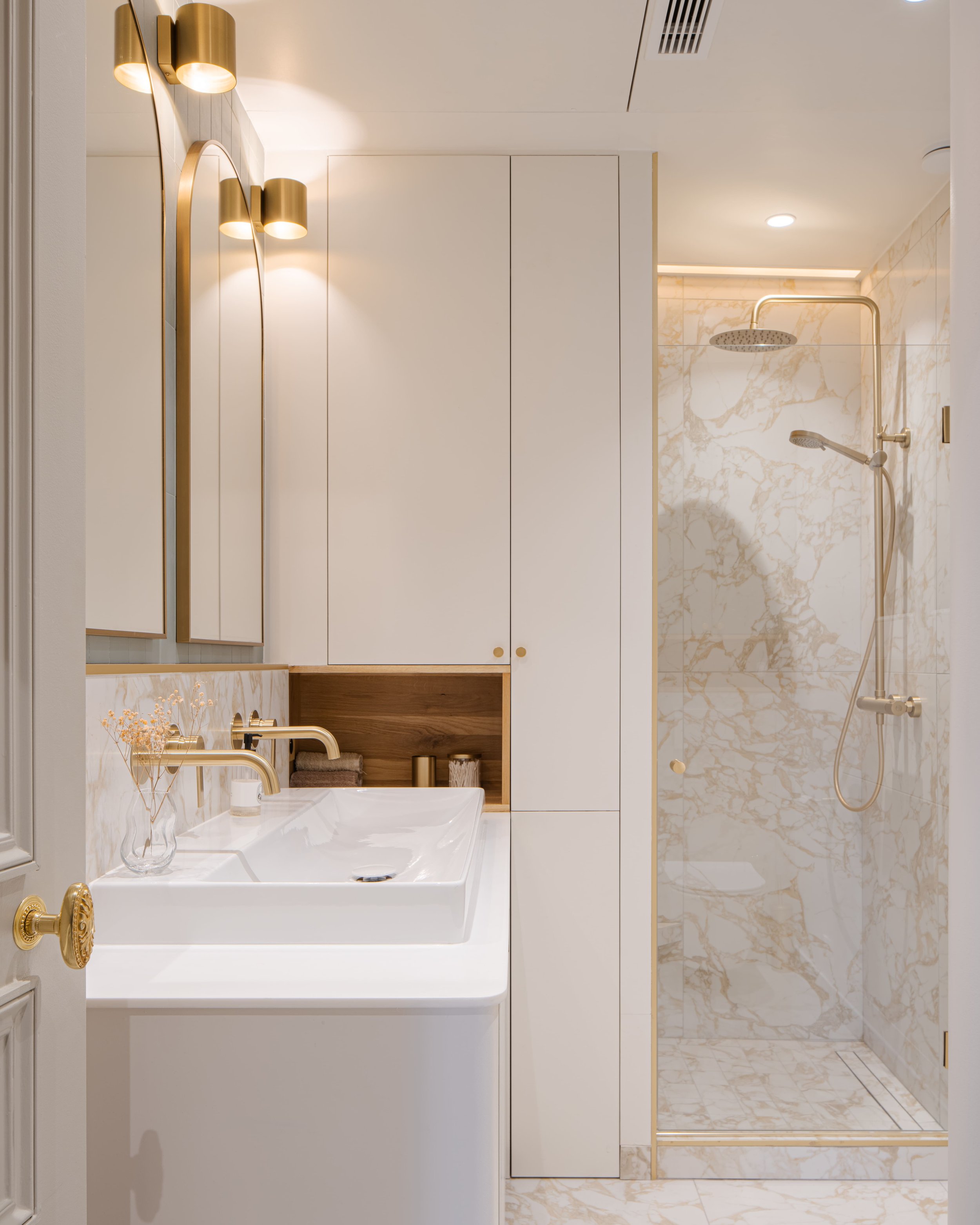 Salle de bain moderne avec un lavabo blanc, un grand miroir, des armoires blanches, et une douche en marbre imitation, accents en or.