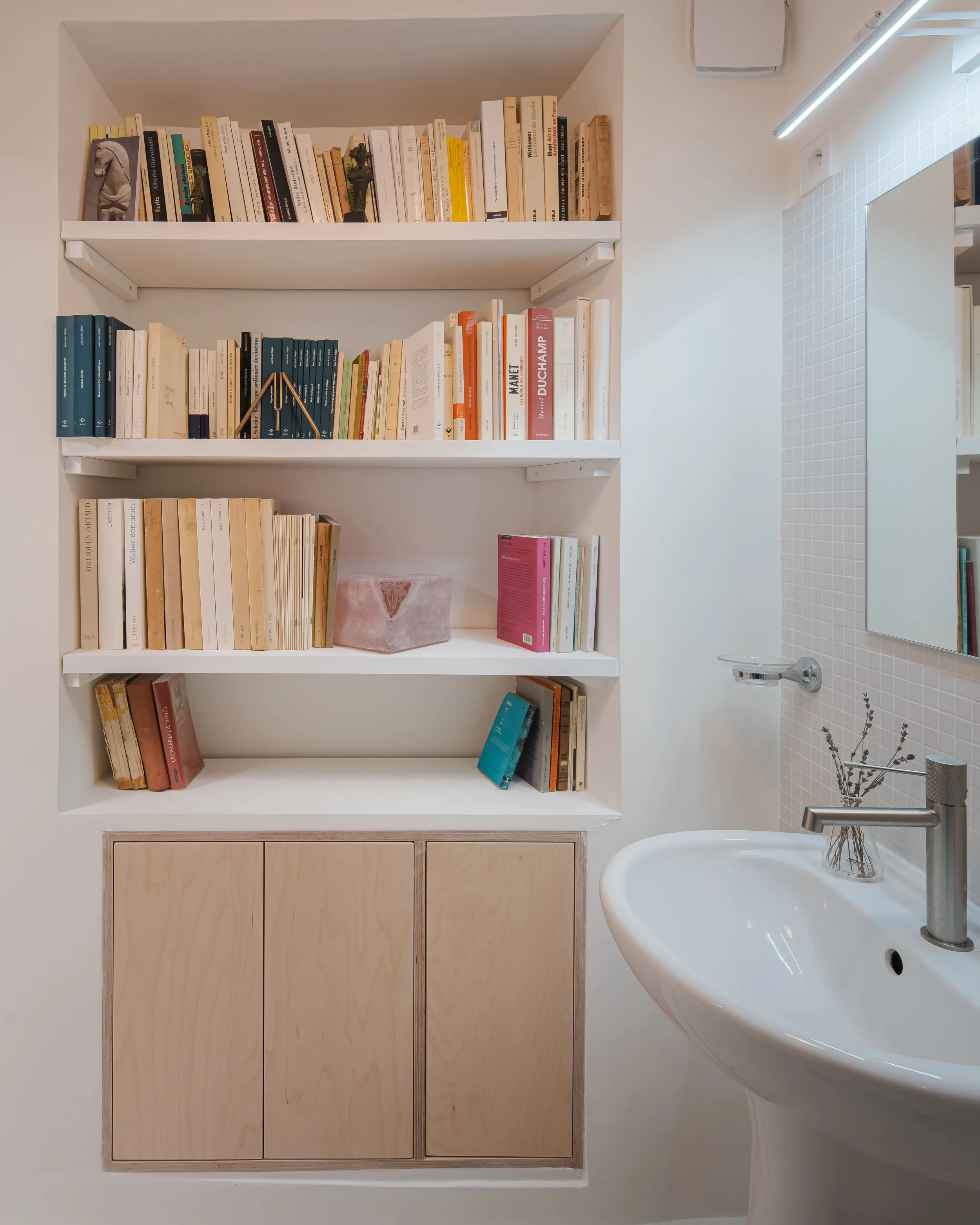 Une étagère blanche avec des livres et un meuble en bois en dessous, située dans une pièce avec un lavabo et un miroir