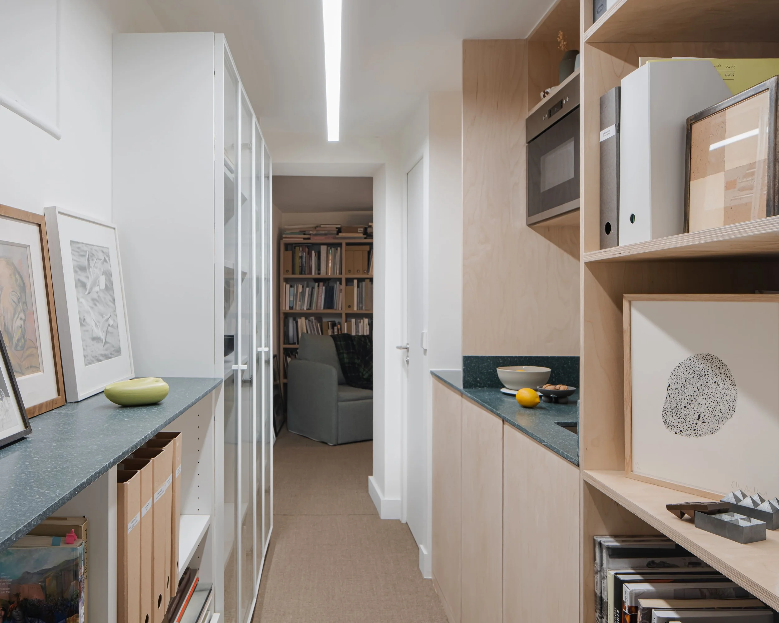 Intérieur d'un appartement moderne avec des étagères, des cadres, des livres et des objets décoratifs, avec un passage menant à un salon avec un fauteuil.
