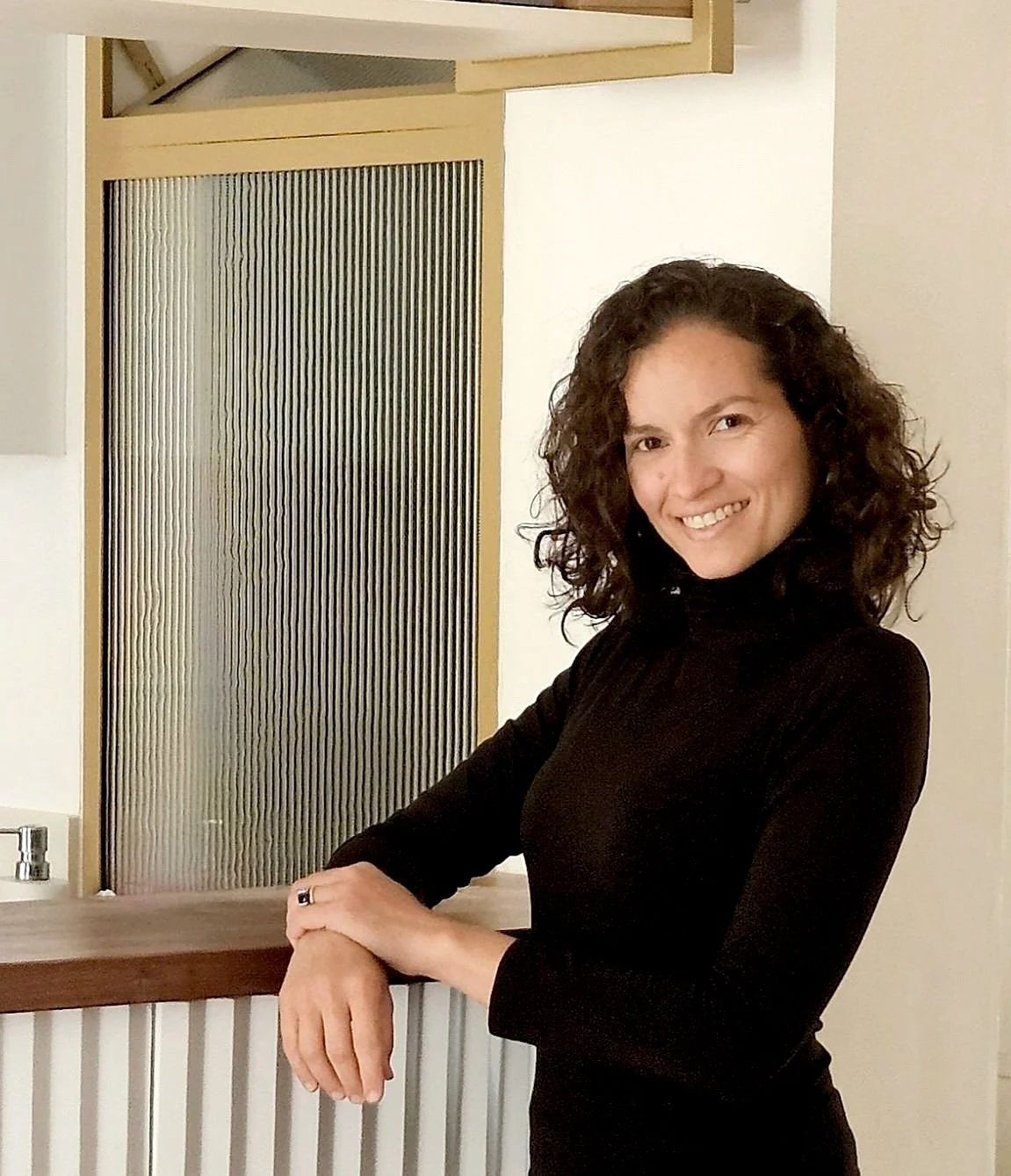 Femme architecte brésilienne souriante avec cheveux bouclés, portant un col roulé noir, posant dans une pièce intérieure près d'un comptoir en bois.