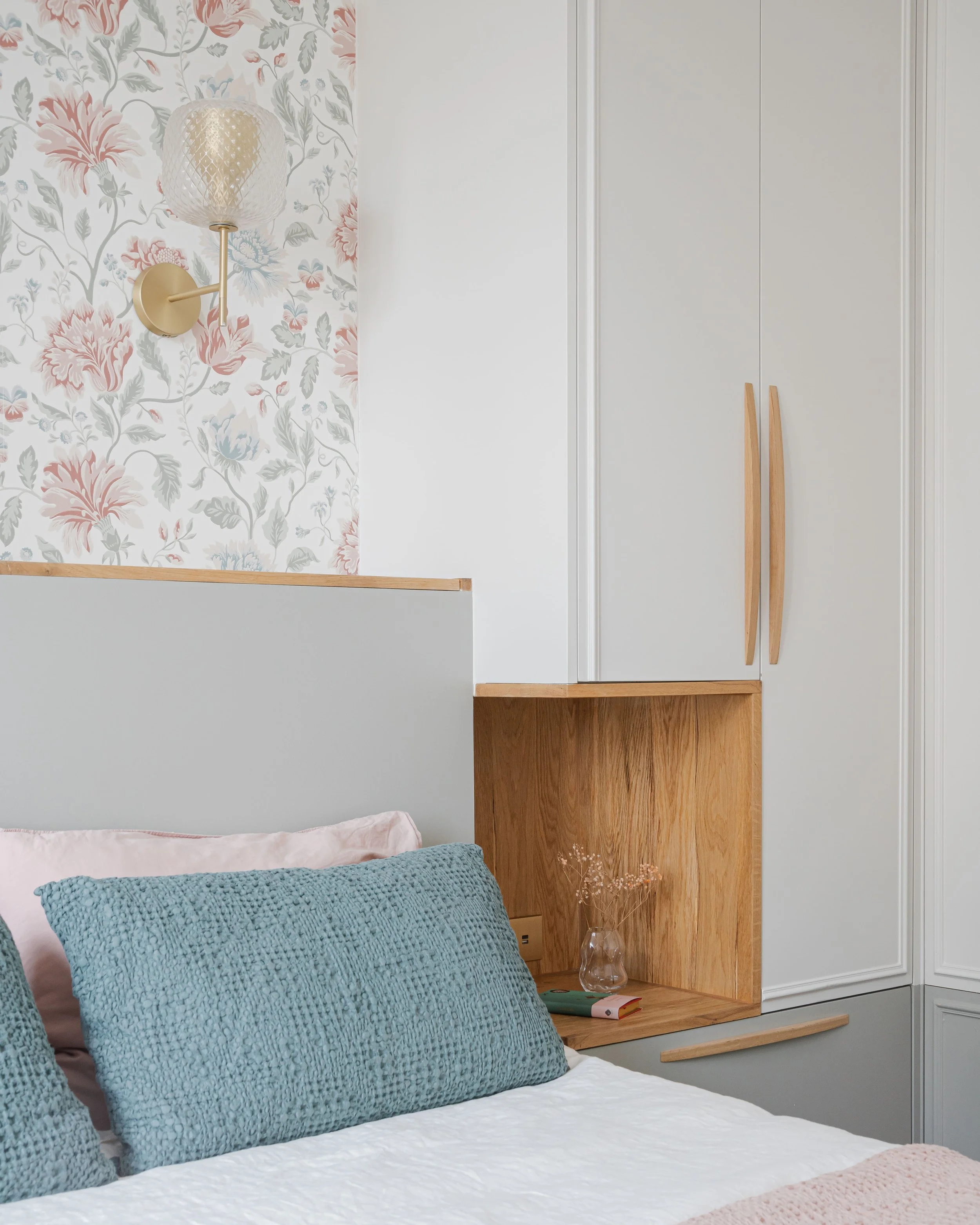 Chambre avec lit blanc, coussins bleus et roses, table de chevet en bois, armoires modernes blanches, lampe murale en forme de boule et fleurs séchées dans un vase en verre.