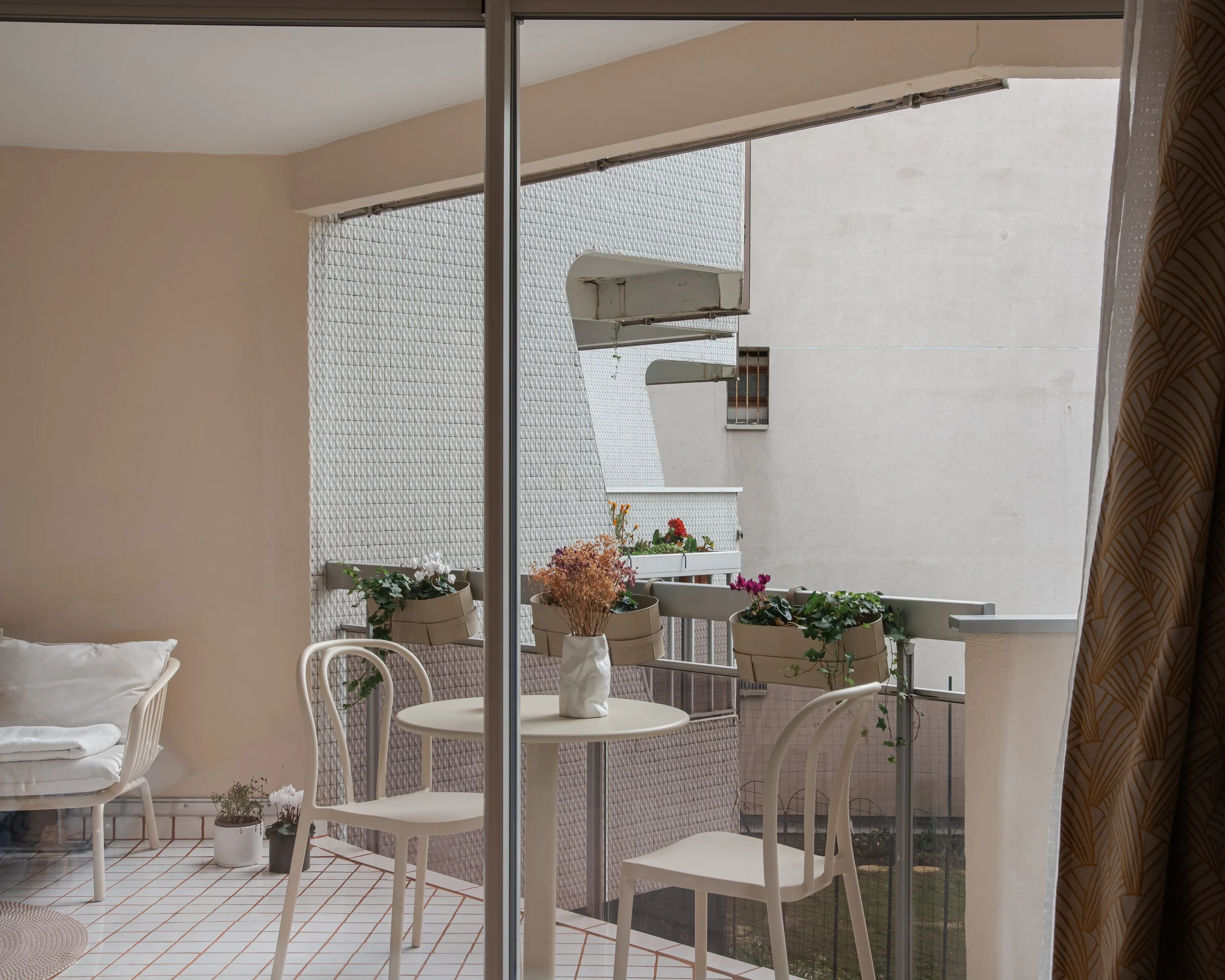Balcon avec table, chaises, et plantes en pot, vue depuis une pièce avec rideau à droite.