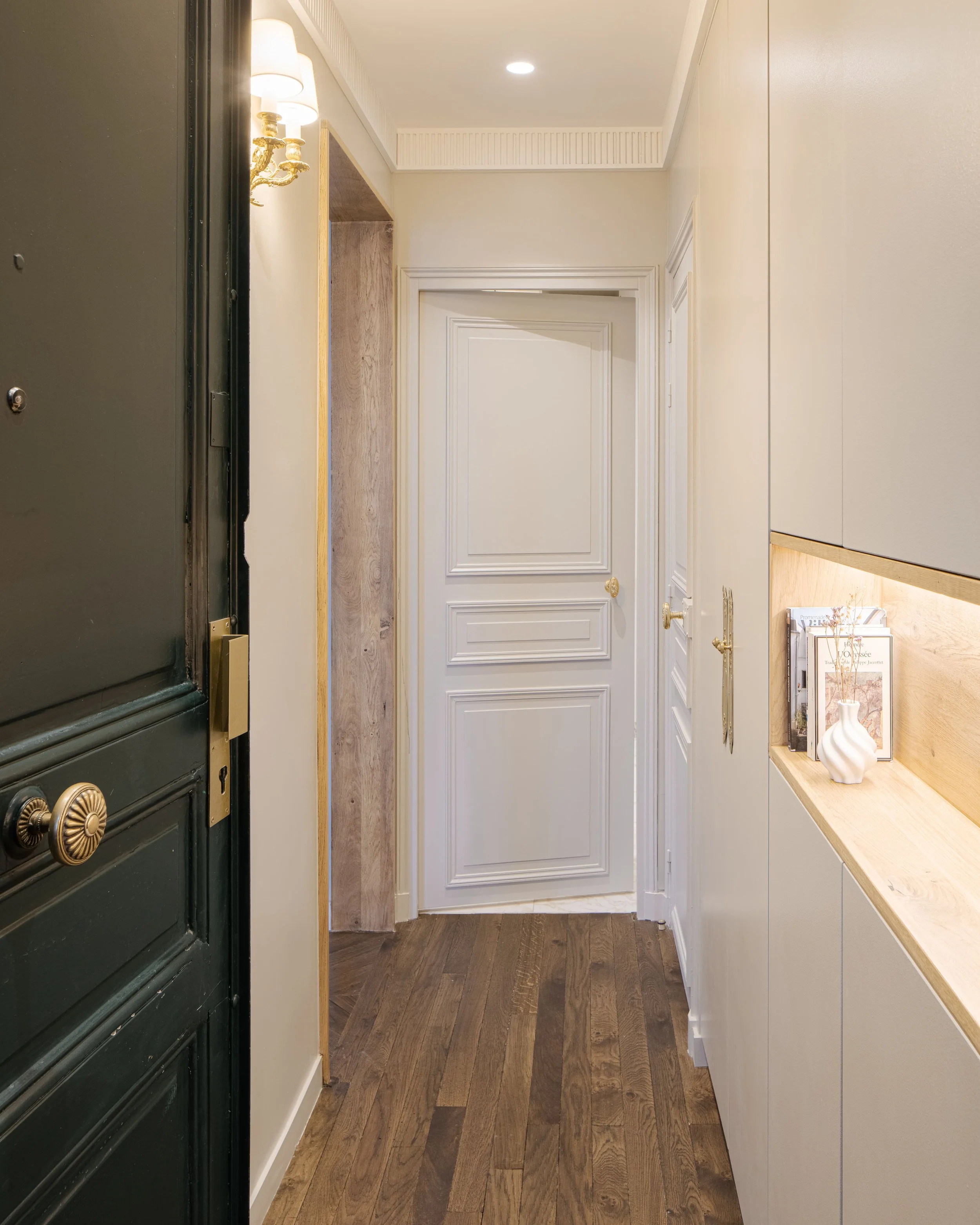Intérieur d'une entrée d'appartement avec un sol en bois, un mur beige, une porte blanche et des éléments décoratifs en bois et céramique.