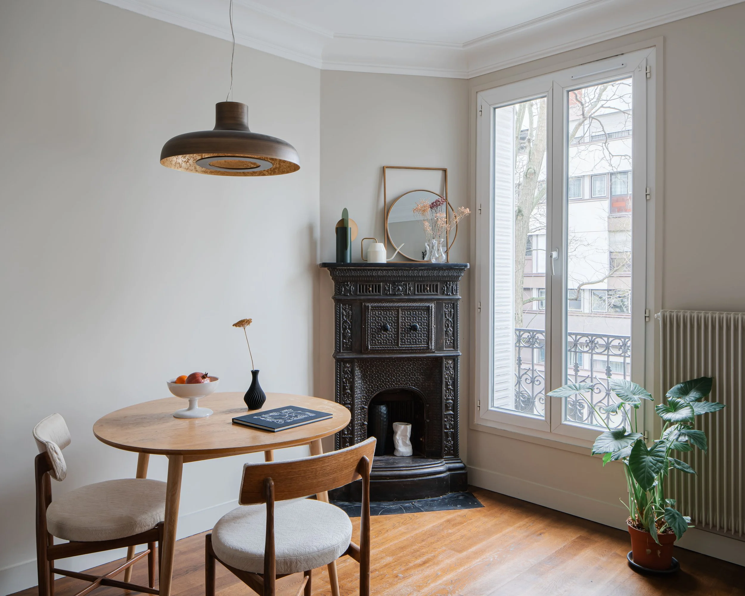Salon avec cheminée noire, grande fenêtre, table ronde en bois, trois chaises, vase avec une fleur, fruits, miroir rond, lampadaire au plafond, plante verte en pot.