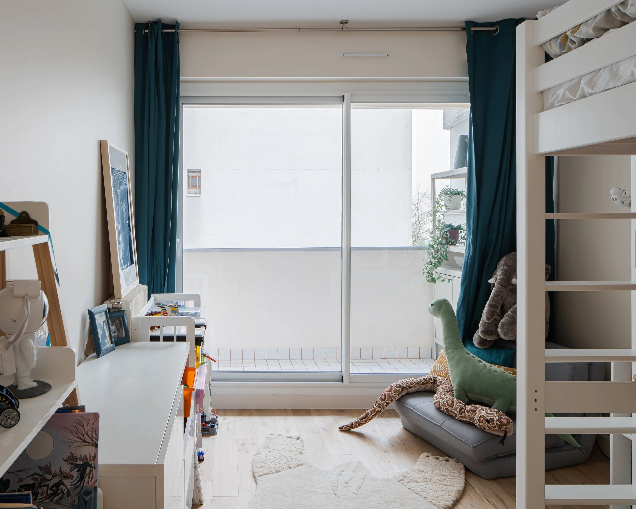 Chambre d'enfant avec un grand balcon, un canapé avec des jouets en forme de dinosaure, une étagère avec des plantes et une cheminée décorative, rideaux bleus, sol en bois, écriture blanche.