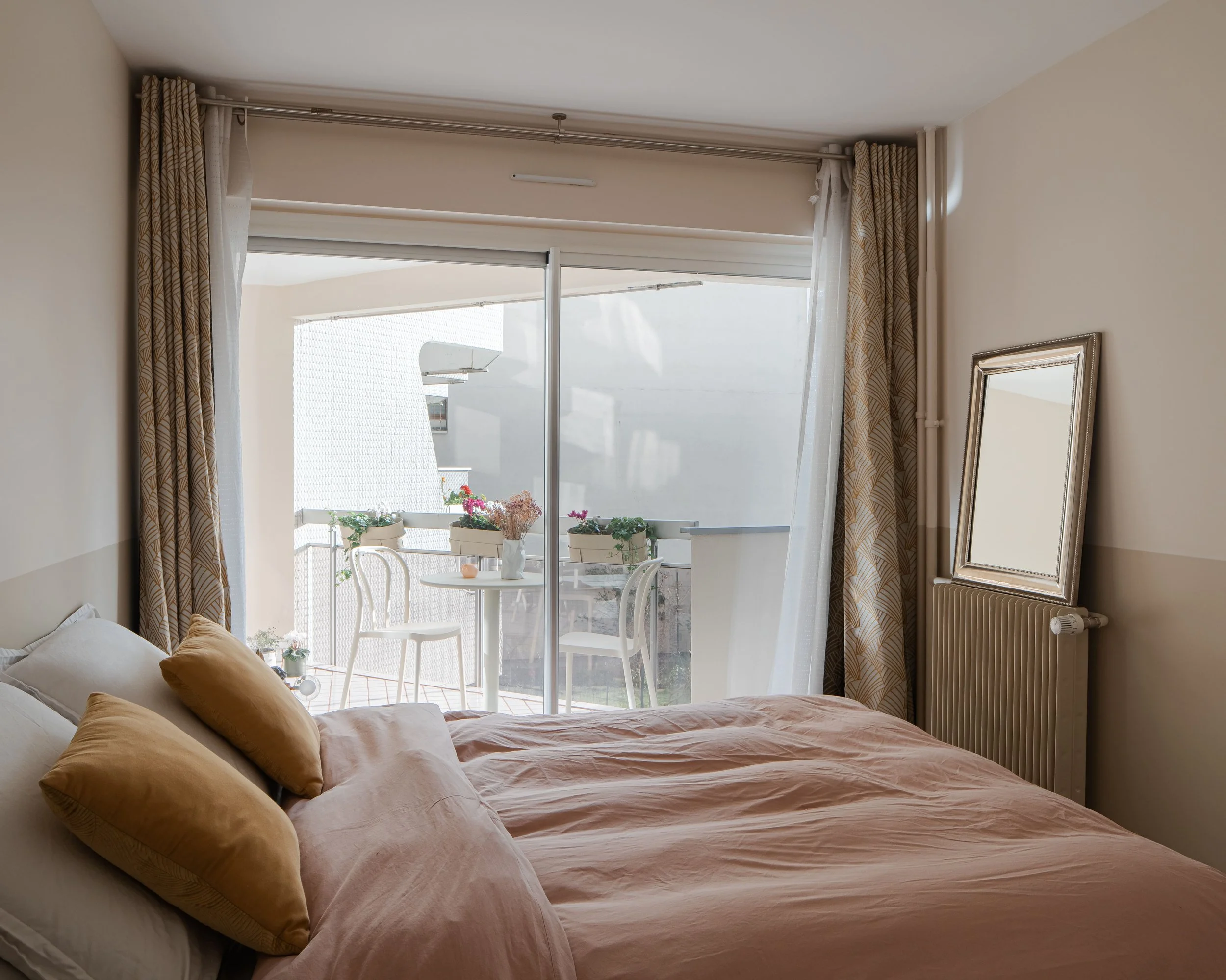 Chambre avec un lit, des oreillers beige, une porte-fenêtre donnant sur un balcon avec une table ronde, deux chaises, des plantes en pots, et un miroir au mur.