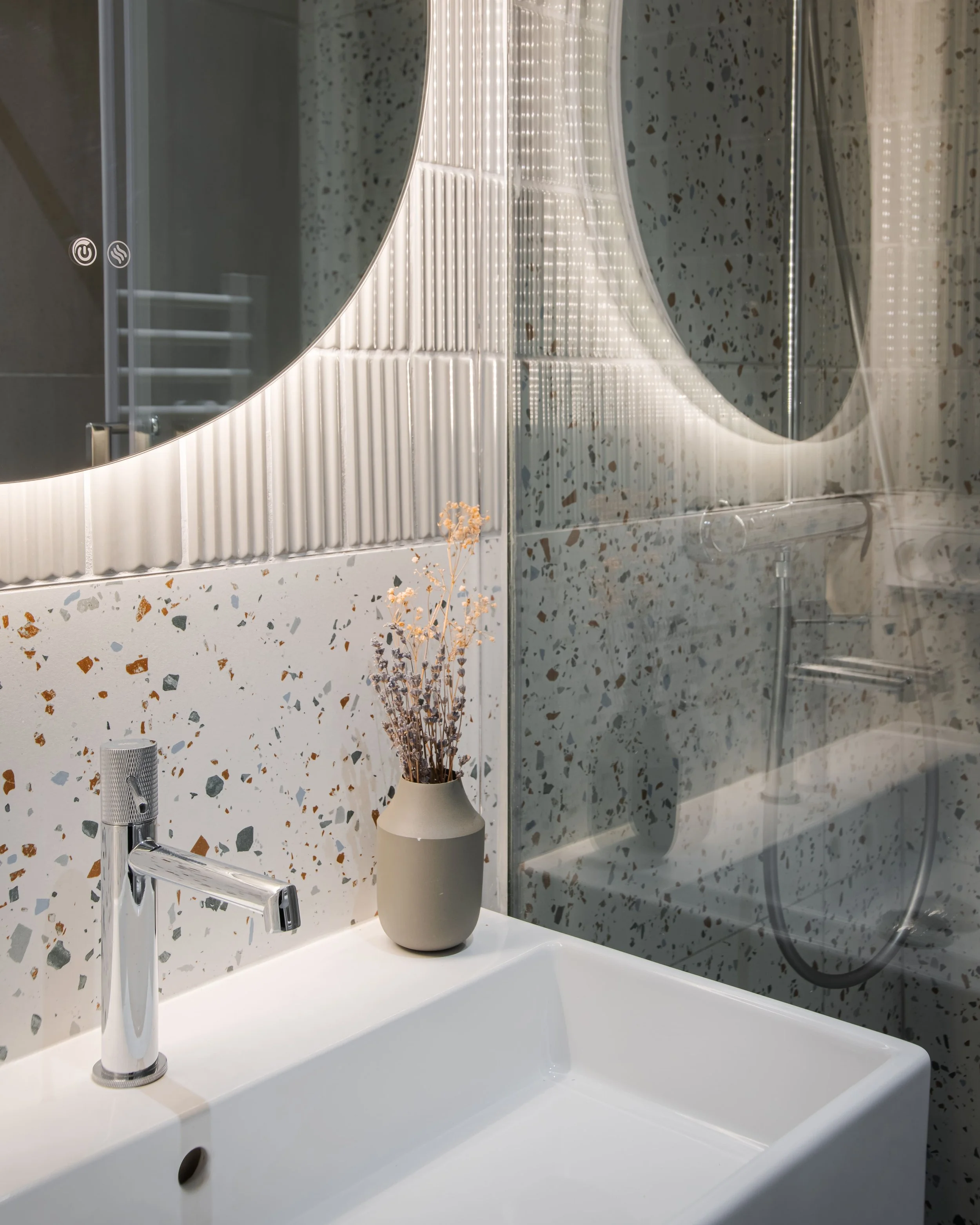 Lavabo moderne avec un miroir rond rétroéclairé, une vase avec des fleurs séchées et une paroi de douche en verre avec un pommeau de douche.