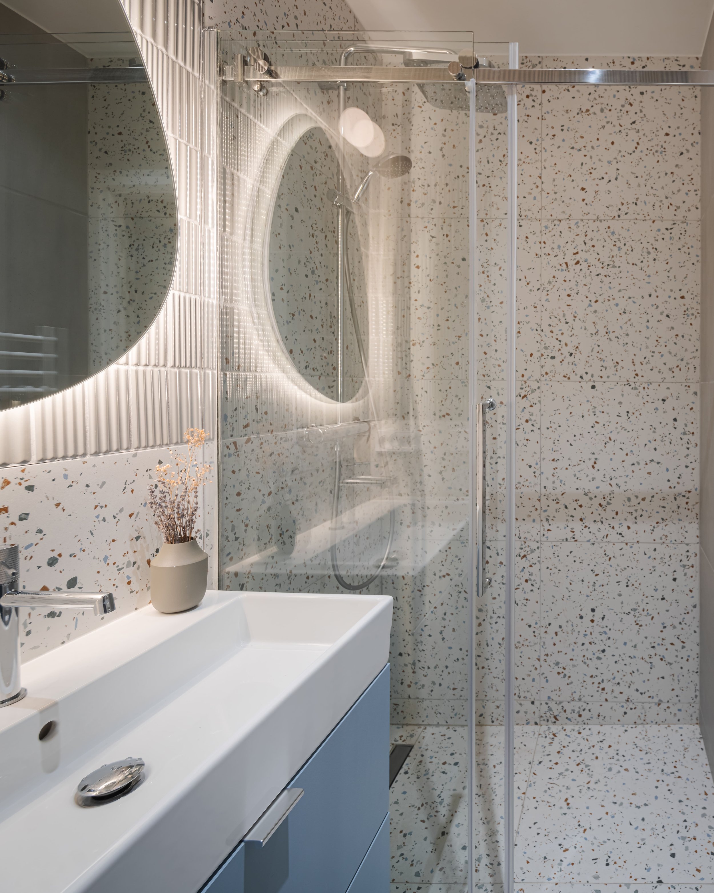 Salle de bain moderne avec miroir rond à LED, douche en verre, lavabo blanc avec vasque rectangulaire, mur en céramique à motifs terrazzo, petite vasque avec fleurs séchées.