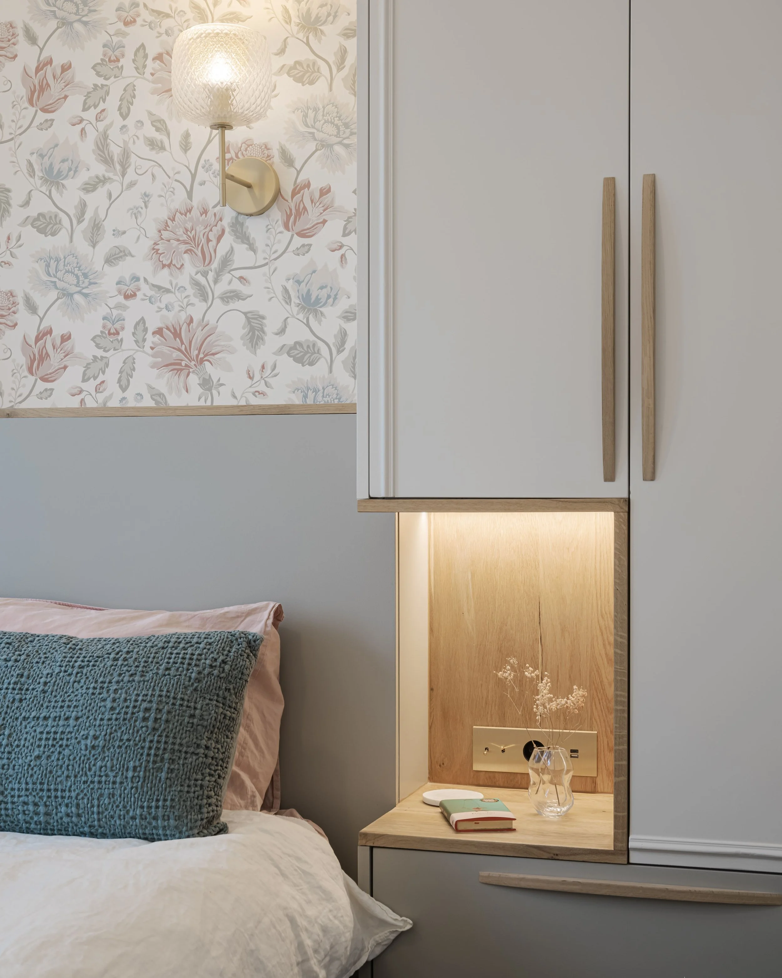 Côté d'un lit avec un coussin bleu et un coussin rose, à côté d'une étagère en bois avec lampe intégrée, une vase avec des fleurs et un livre