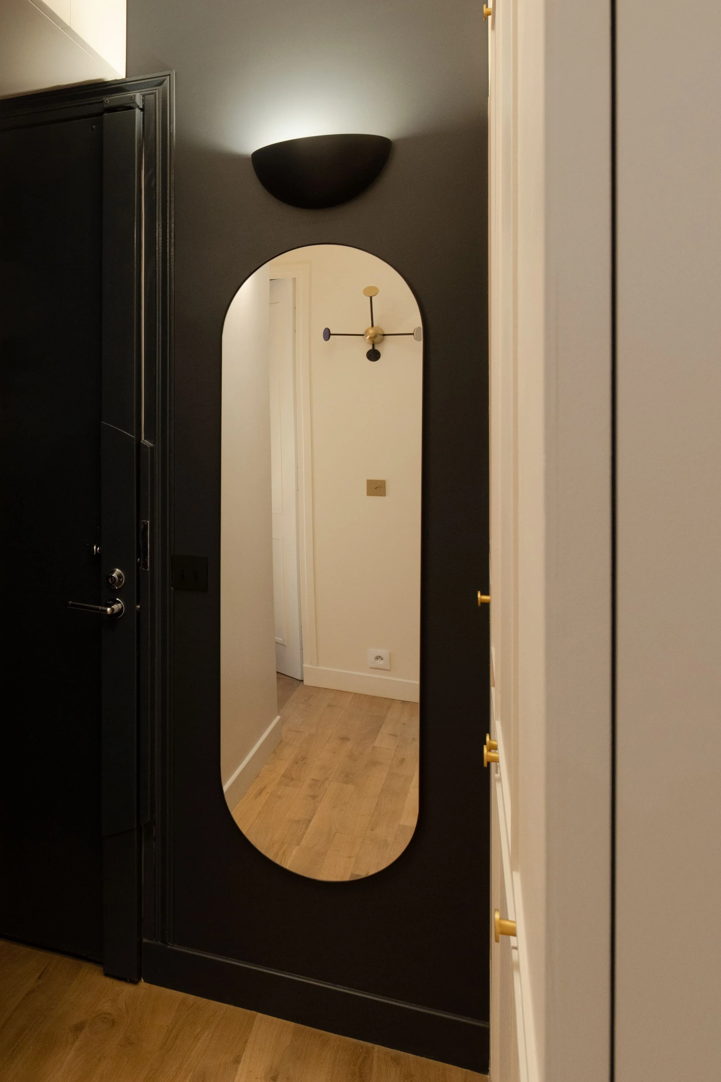 Un miroir ovale fixé sur un mur noir, reflétant une porte blanche dans un intérieur moderne avec un sol en bois. Au-dessus du miroir, un luminaire mural noir et une horloge murale avec bras bleus, une aiguille noire et une couleur dorée.