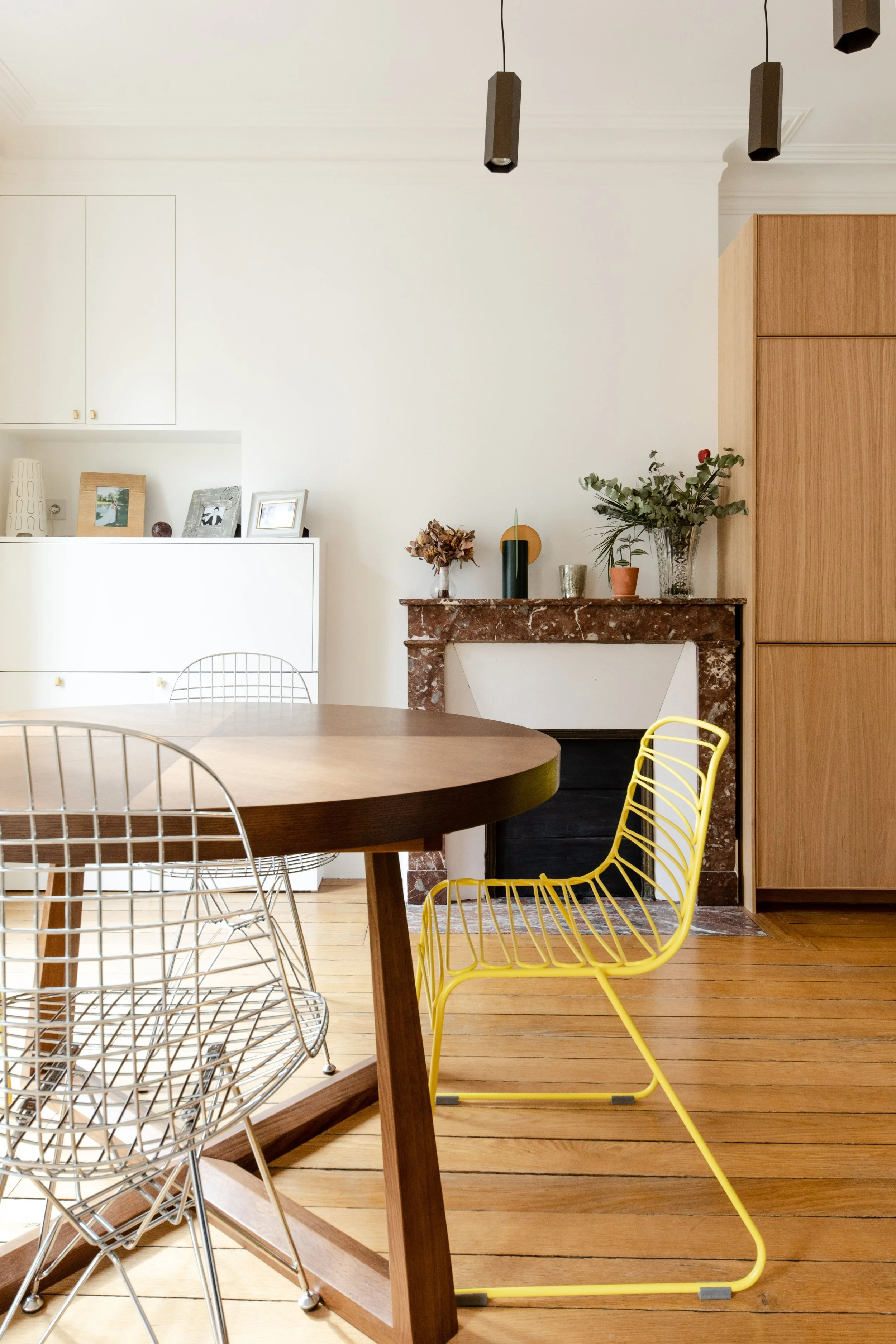 Intérieur d'une salle à manger avec une table en bois, chaises en métal, cheminée en marbre décorée avec des plantes et des cadres photo, plafond avec deux lampes suspendues