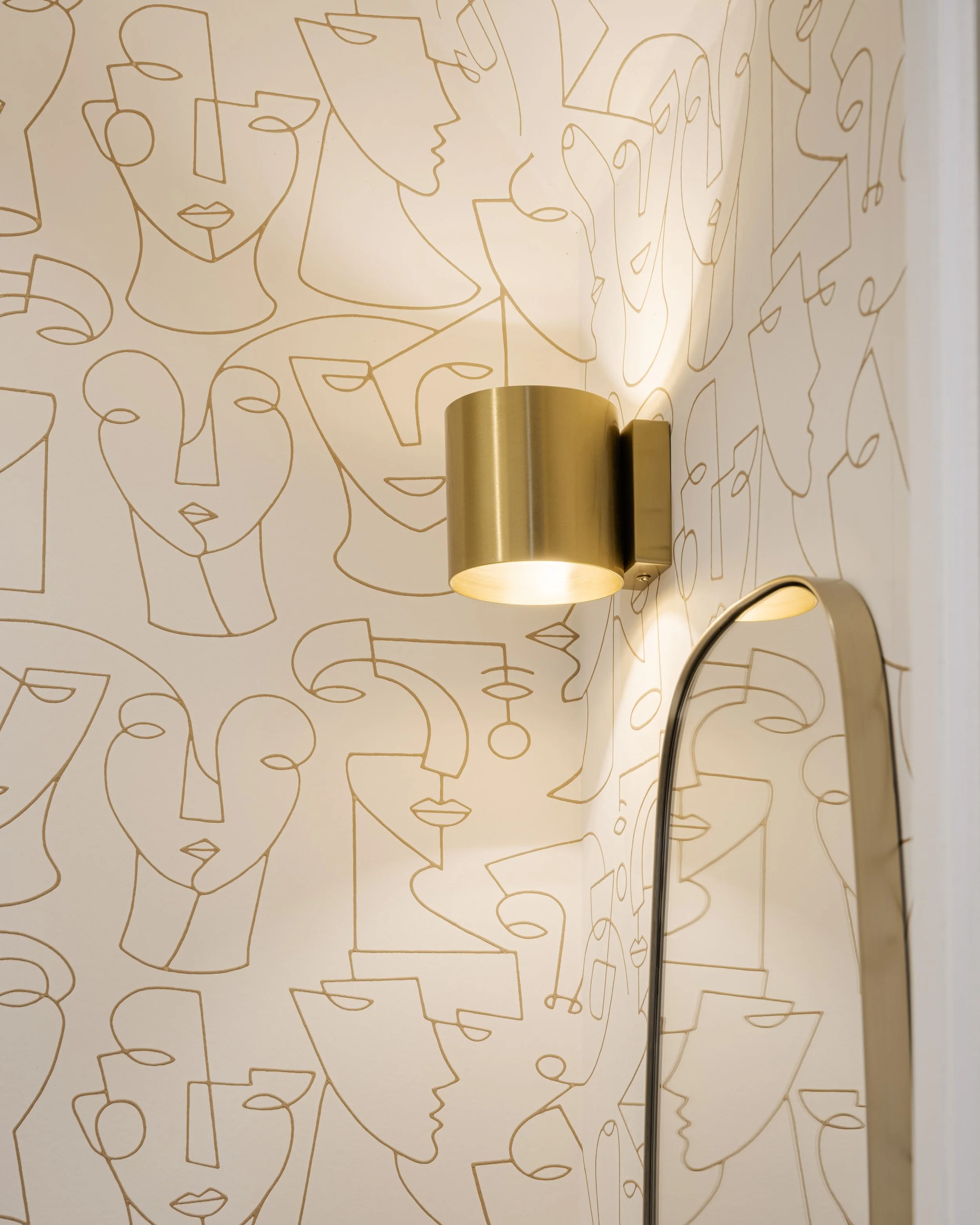 Mural avec dessins stylisés de visages féminins en lignes dorées sur un mur blanc, avec une lampe murale en or et un miroir incurvé.