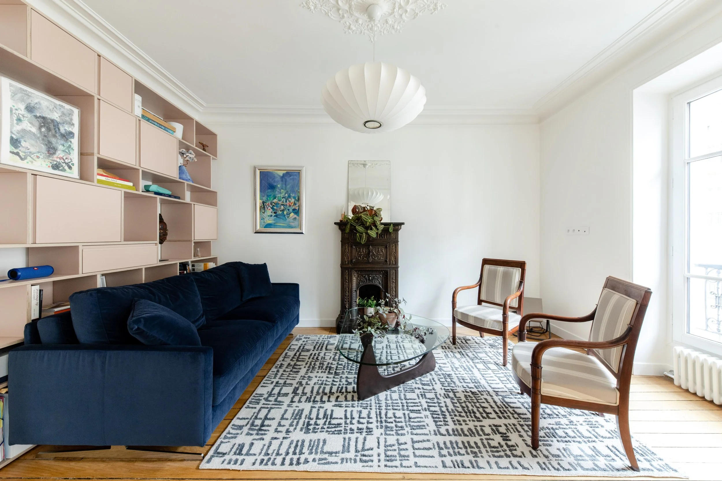 Salon avec canapé en velours bleu foncé, deux fauteuils en bois et tissu beige, table en verre, cheminée ancienne en bois, étagère murale rose, tapis noir et blanc, peinture colorée, miroir, plantes en décoration.