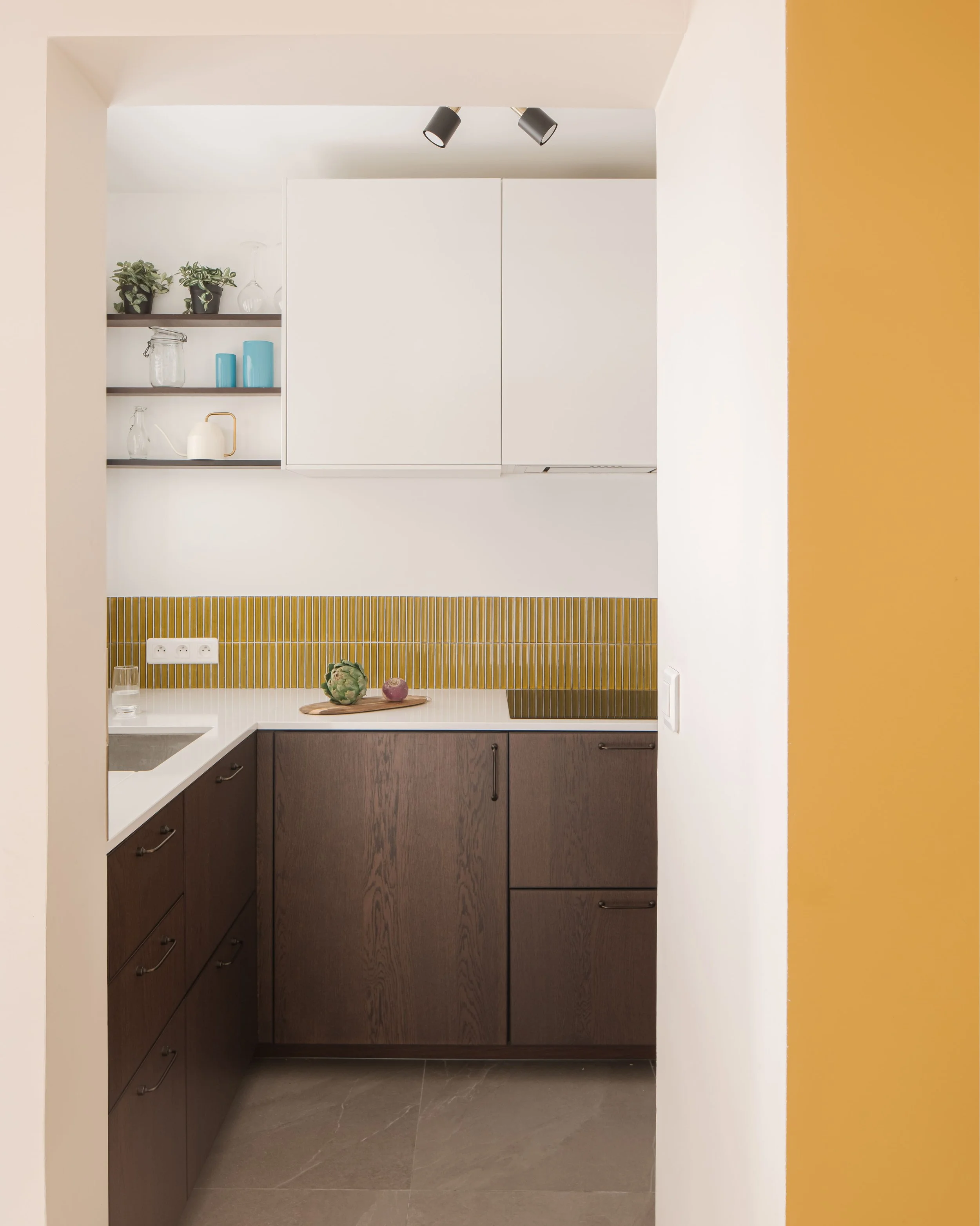 Petite cuisine moderne avec moulures blanches, plan de travail blanc et meubles en bois foncé. Étagères avec plantes et vaisselle, derrière une ouverture avec murs blanc et jaune moutarde.