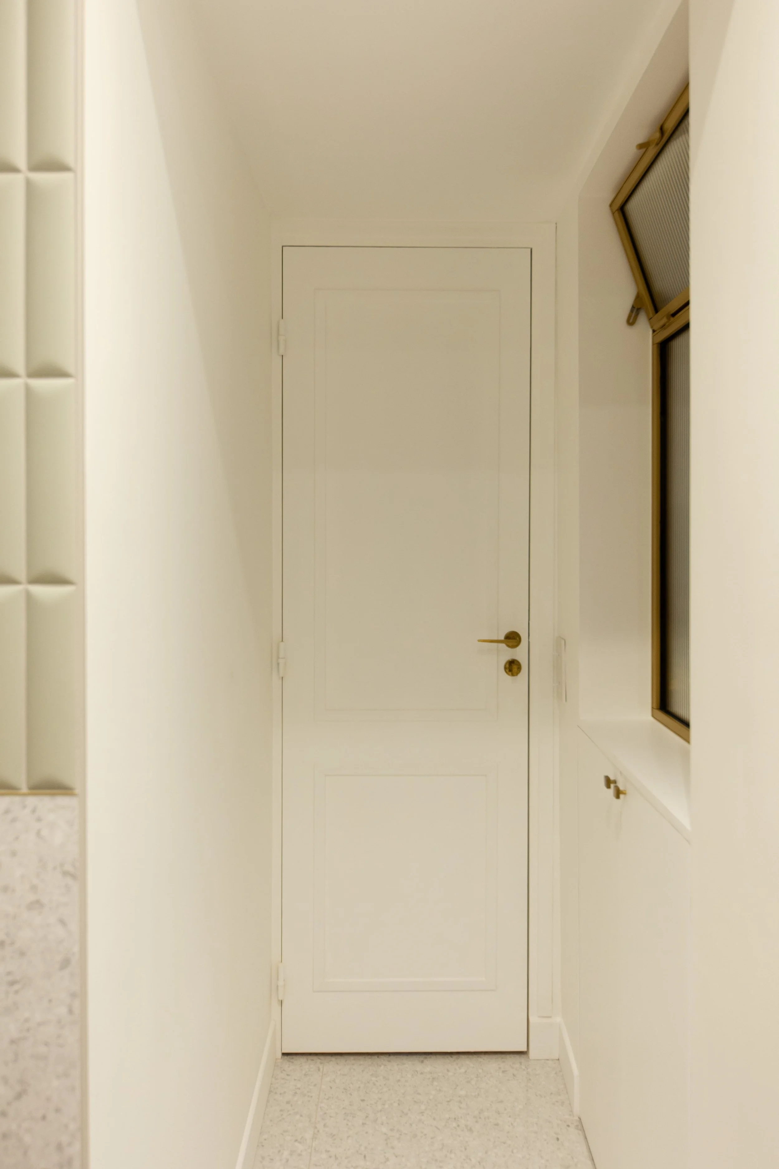 Une porte blanche dans un couloir lumineux avec un mur beige à gauche et une fenêtre avec volet roulant doré à droite.