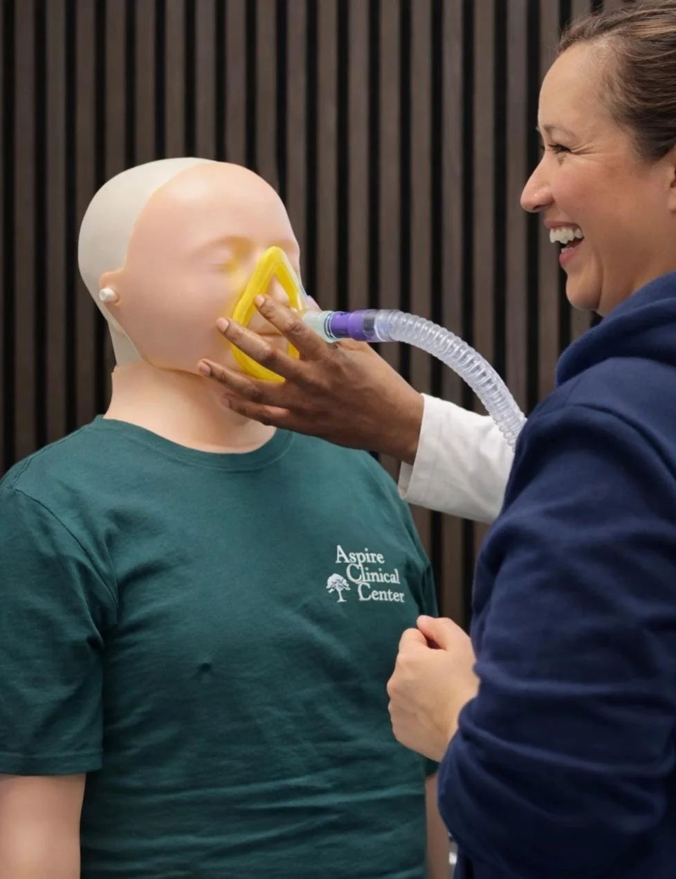 Leaner delivering cough assist to sim manikin face whilst another learner is smiling