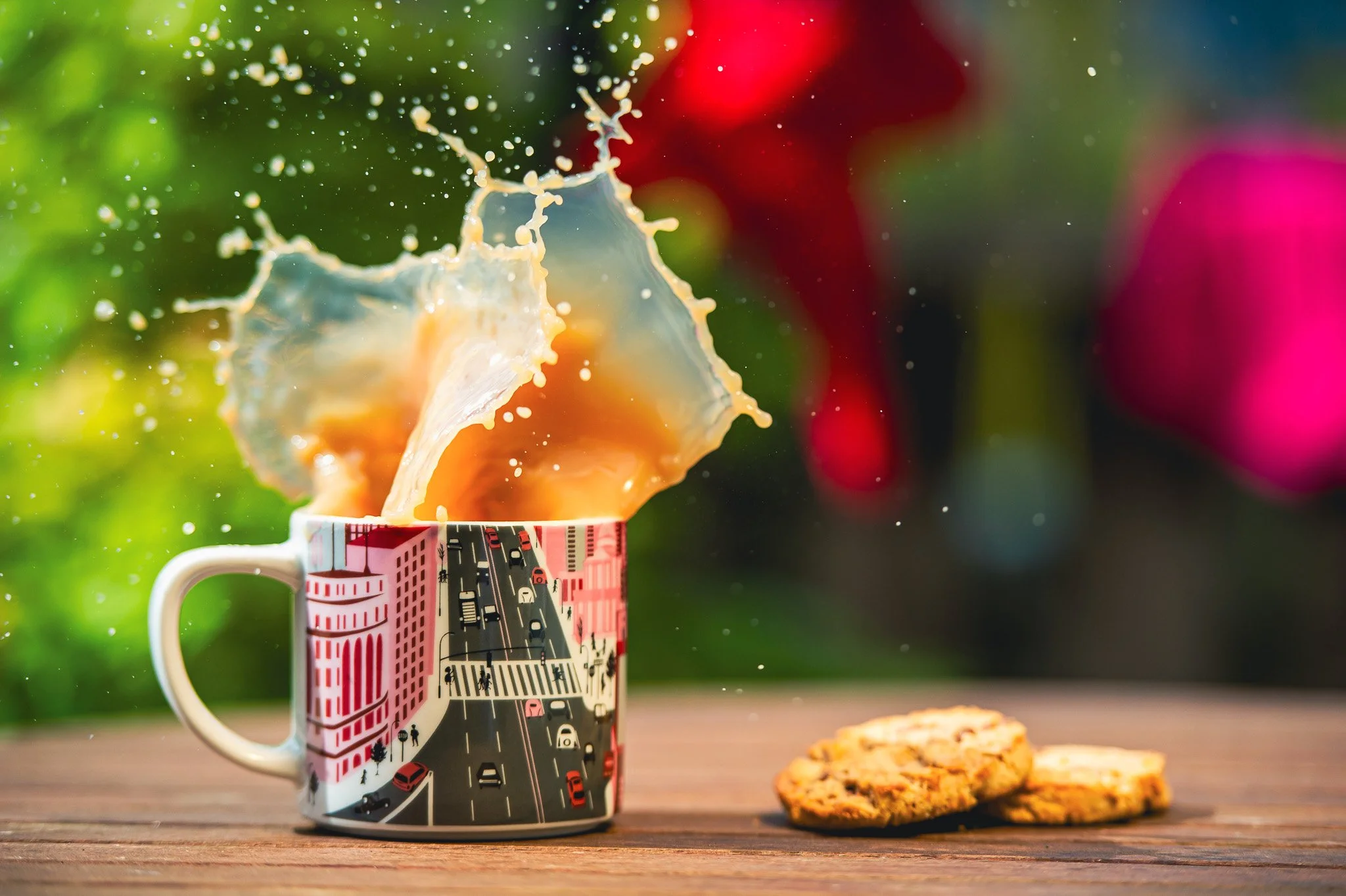 Coffee mug with city street design on it, with coffee spilling over the top into the air, and two cookies on the wooden surface nearby, against a colorful blurred background.