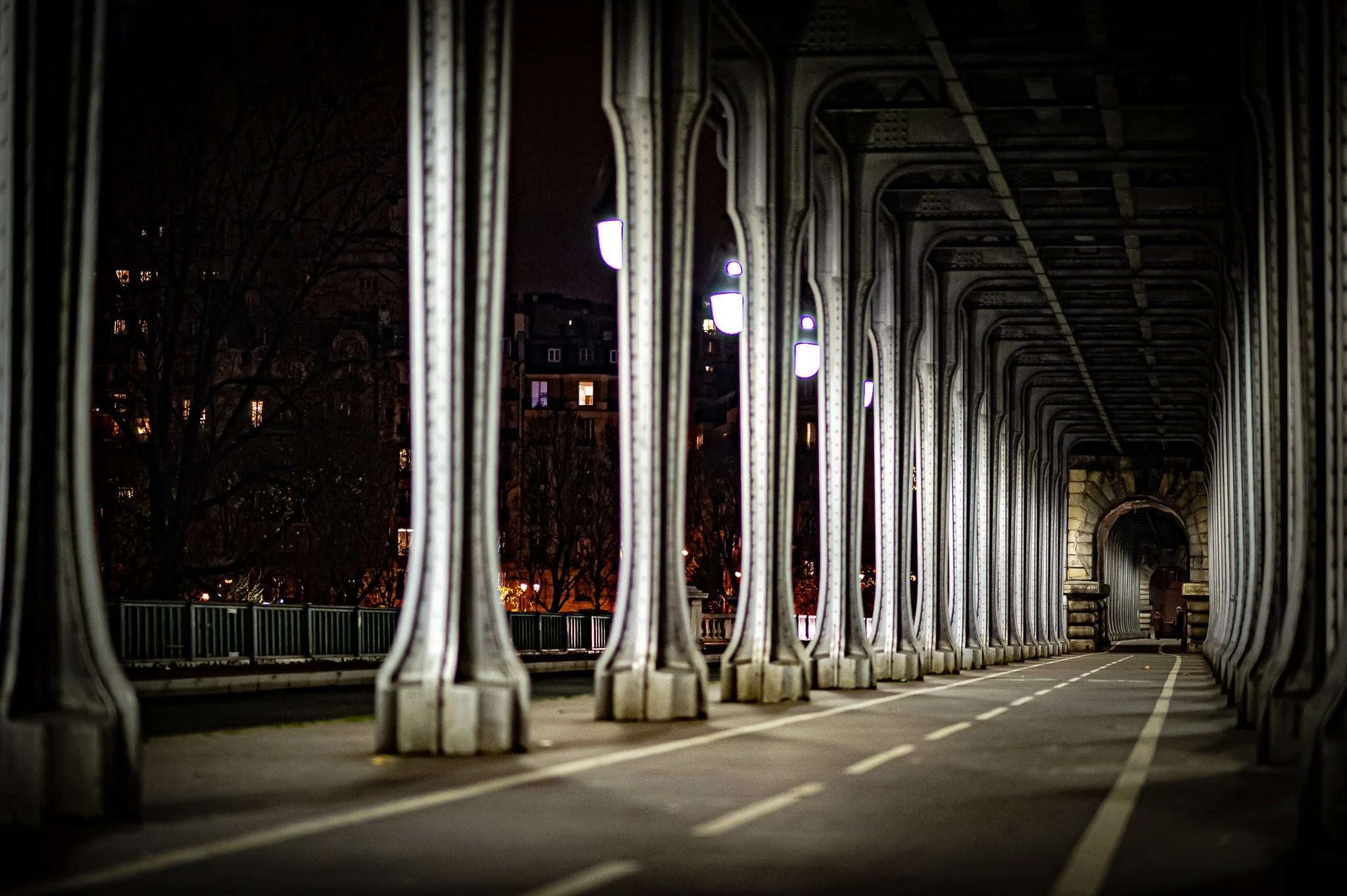 Night view of a pedestrian walkway under a bridge with metal columns, lit by streetlights, with buildings and trees in the background.