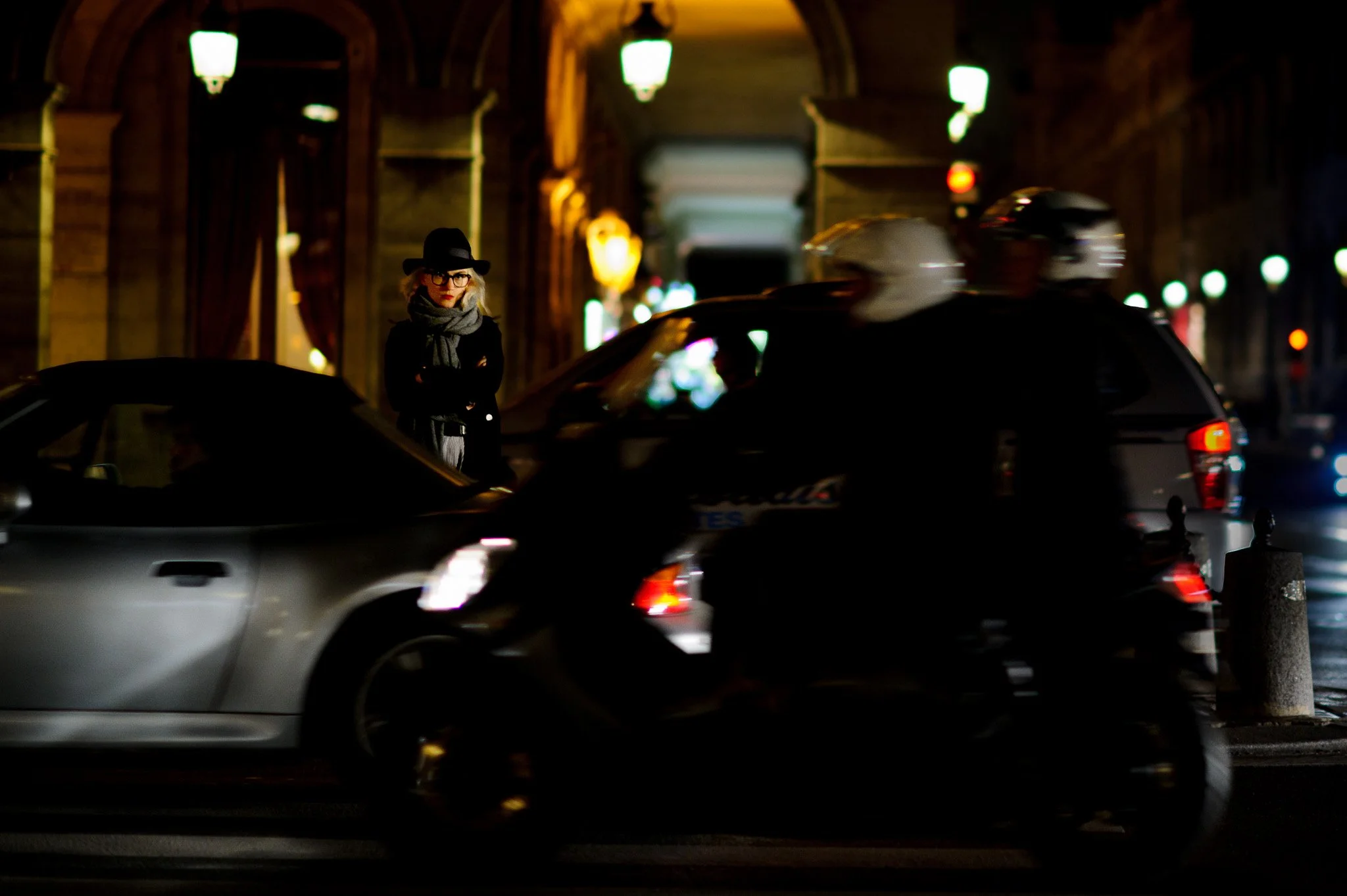 A woman in a black hat, glasses, and scarf standing on a city street at night, with moving vehicles including a silver car and a motorcycle passing by.