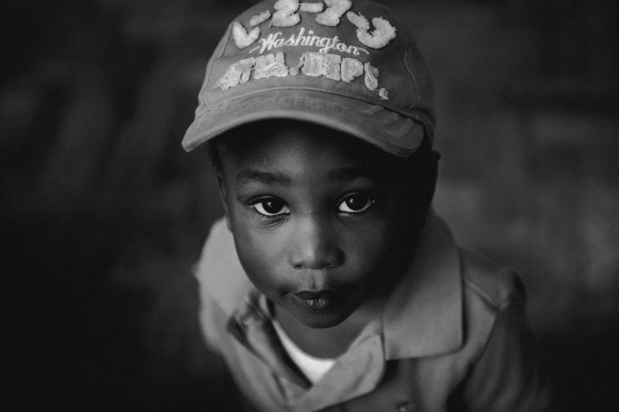 A young boy looking up at the camera, wearing a baseball cap and a collared shirt, in black and white.