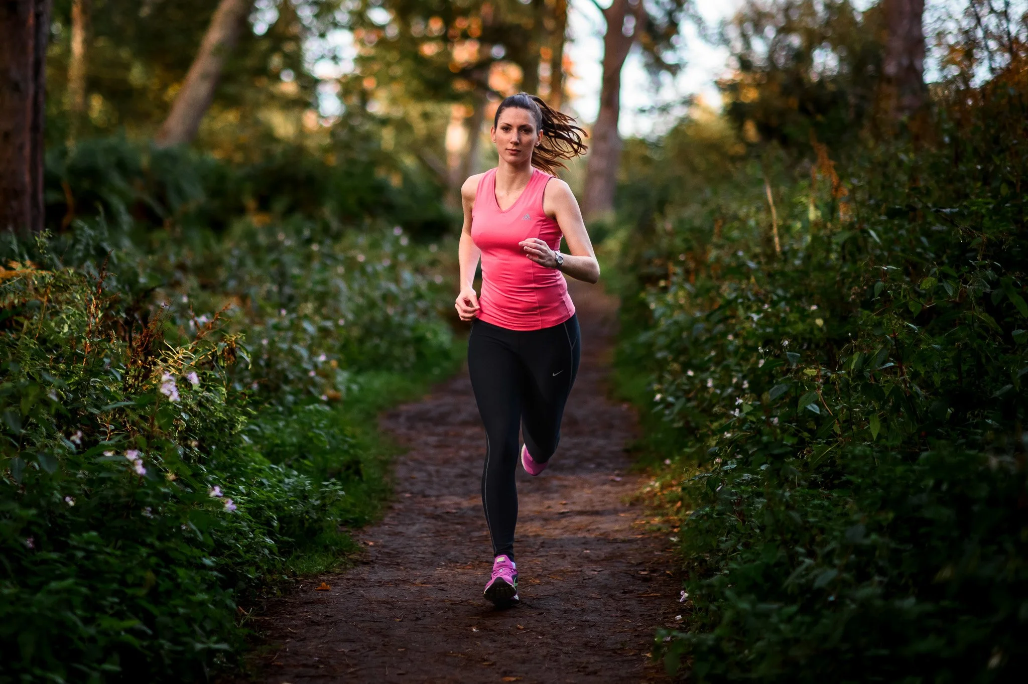 A woman running on a dirt trail in a forested area during the daytime, wearing a pink tank top, black leggings, and pink running shoes.