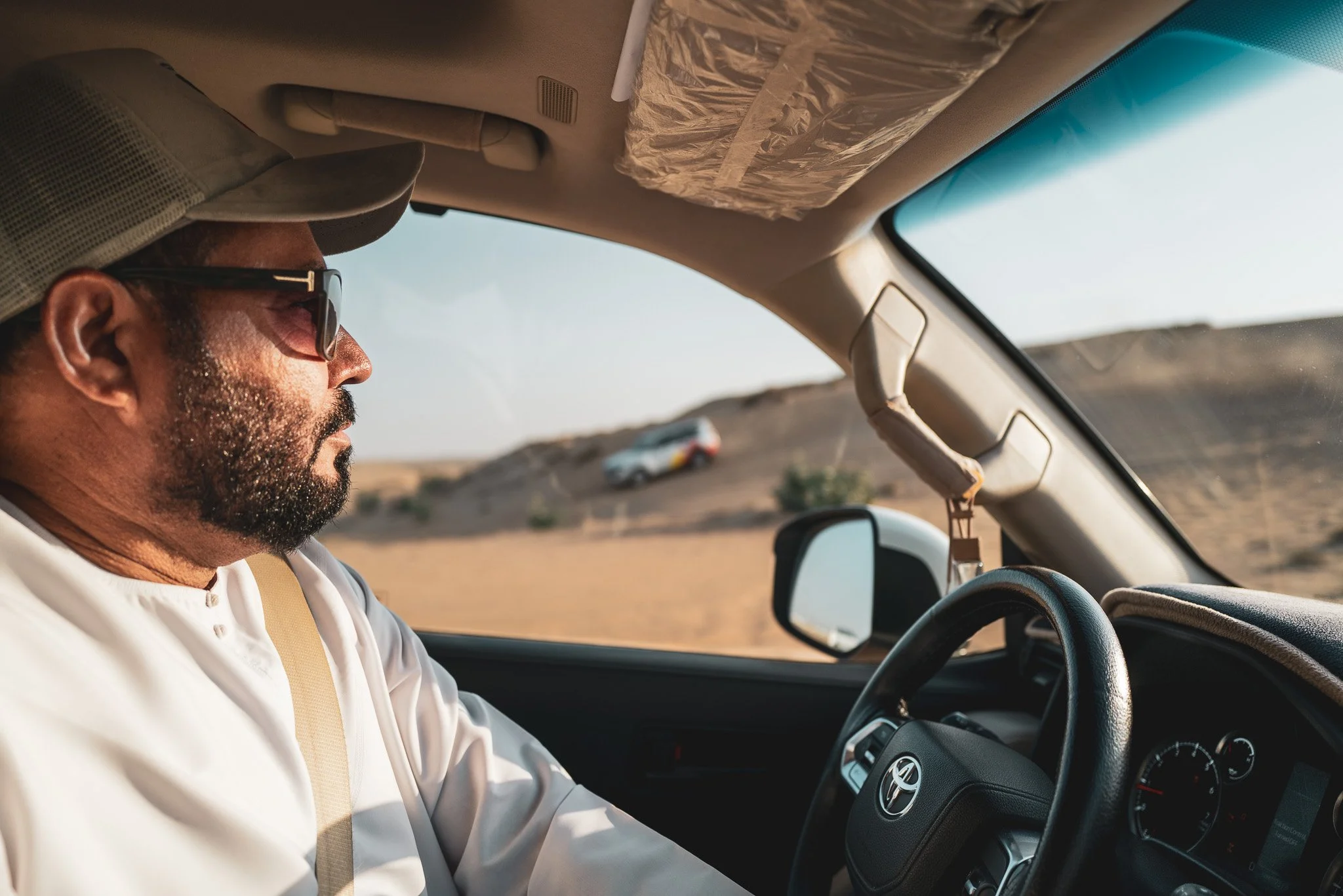 Man driving a Toyota vehicle through a desert landscape with a car on a hill in the distance.