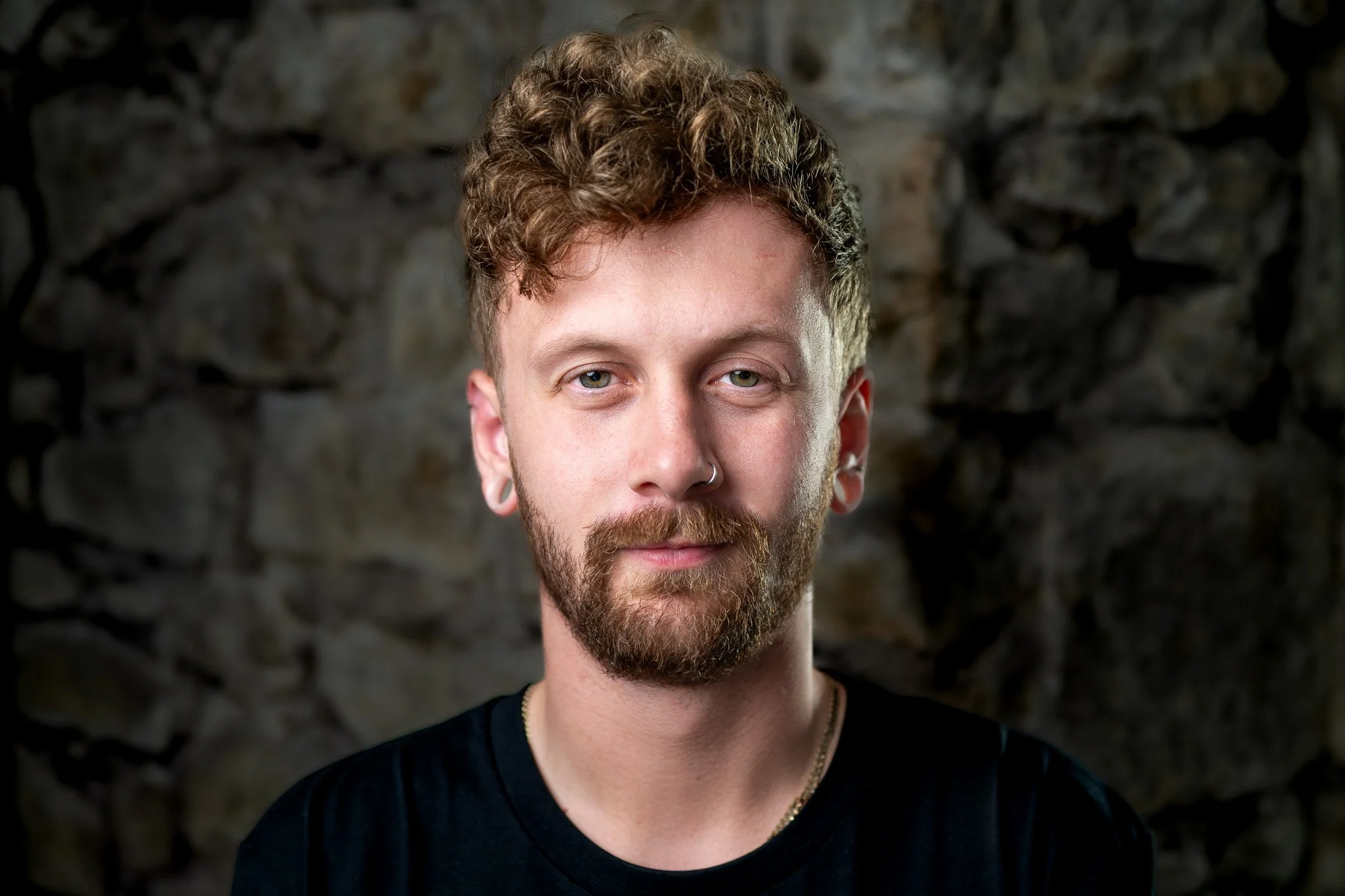 Portrait of a young man with curly hair and beard, wearing a black shirt, standing against a stone wall.