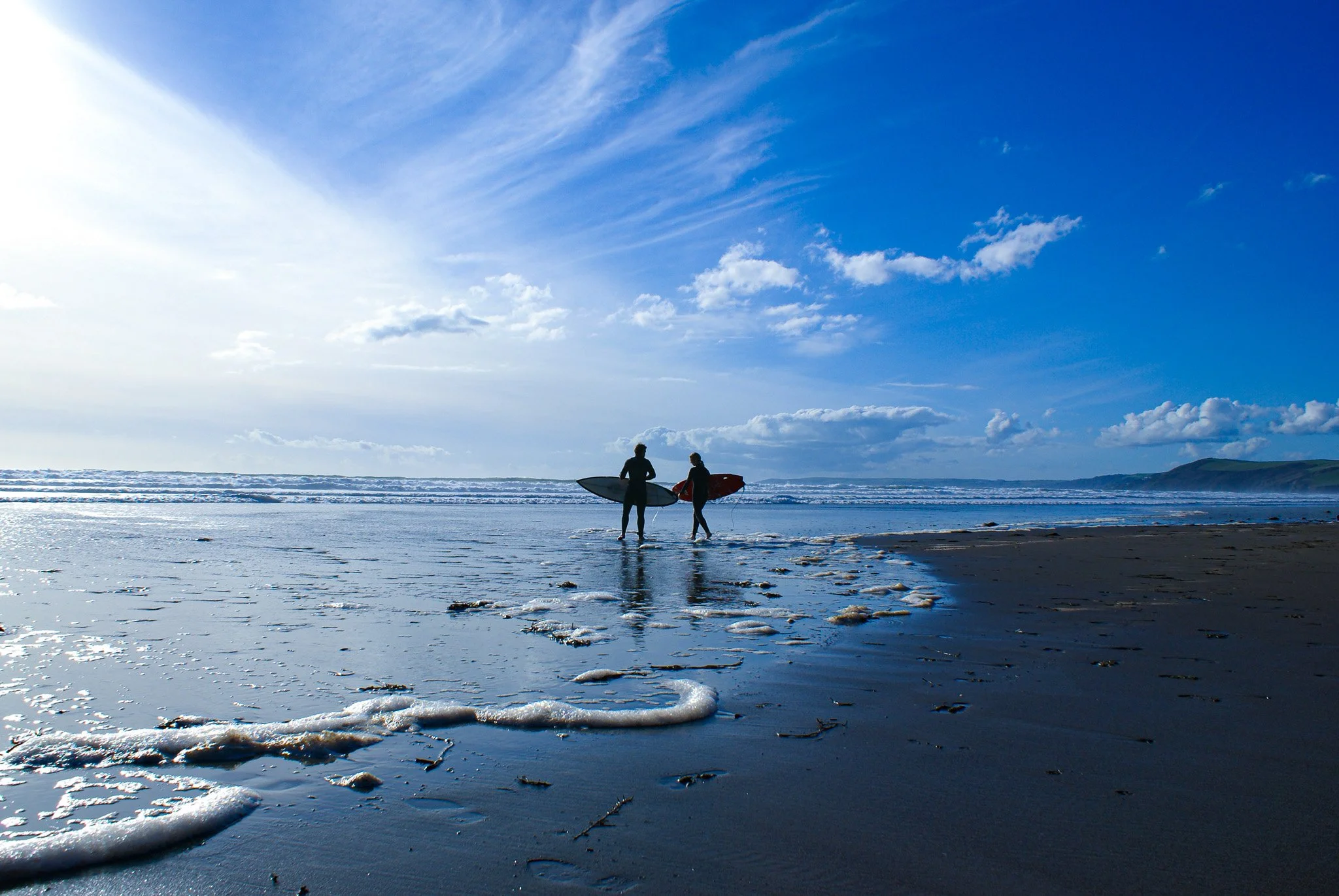 Two surfers walking on the beach carrying surfboards, with the ocean and a blue sky in the background.