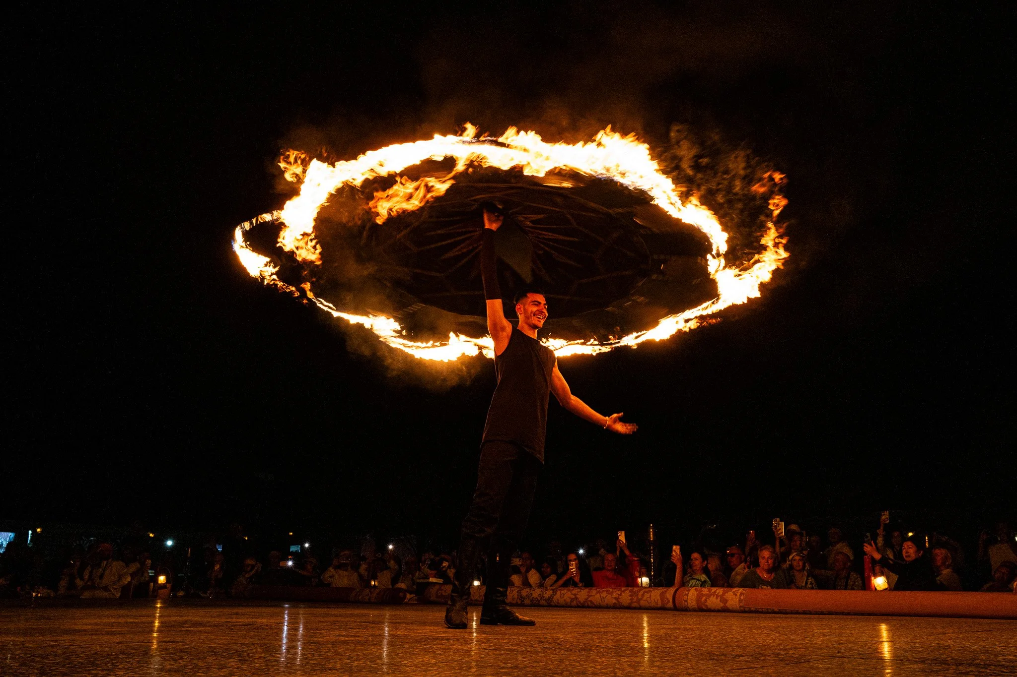 A fire performer spins a flaming ring at night while an audience watches and takes photos nearby.