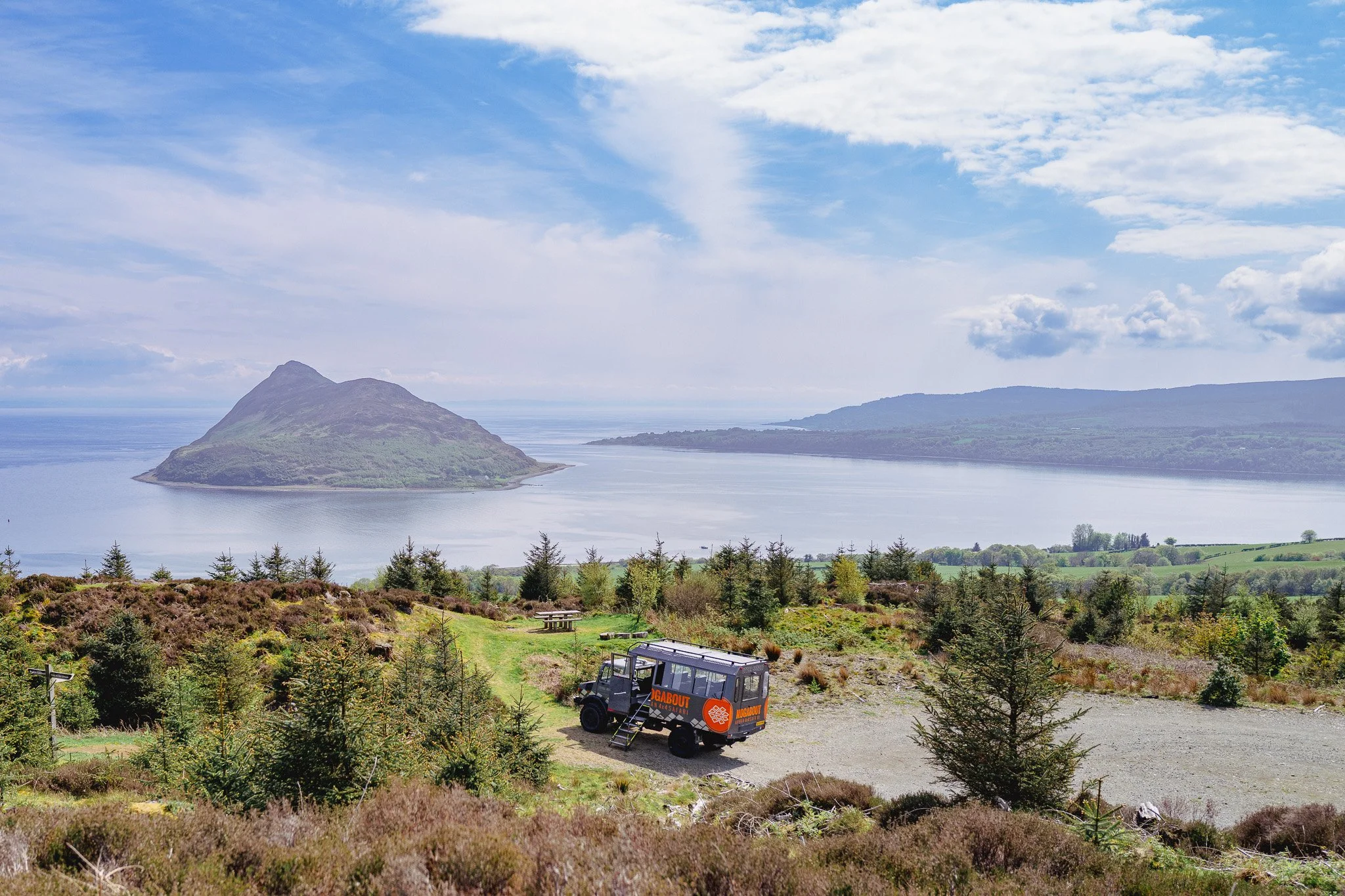A scenic landscape with a large lake, a mountainous island in the background, and a grassy foreground with small trees. A black tour vehicle with orange and white signage is parked on a gravel area near the trees. There are picnic tables in the dista