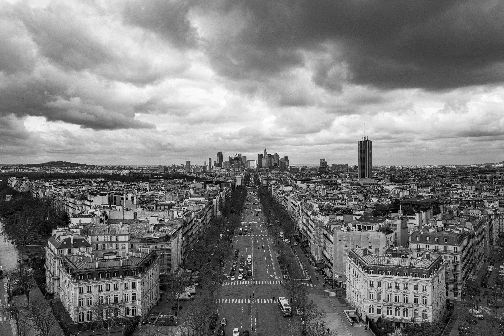 A black-and-white aerial view of a city with a wide central avenue, tall buildings in the distance, and cloudy skies overhead.