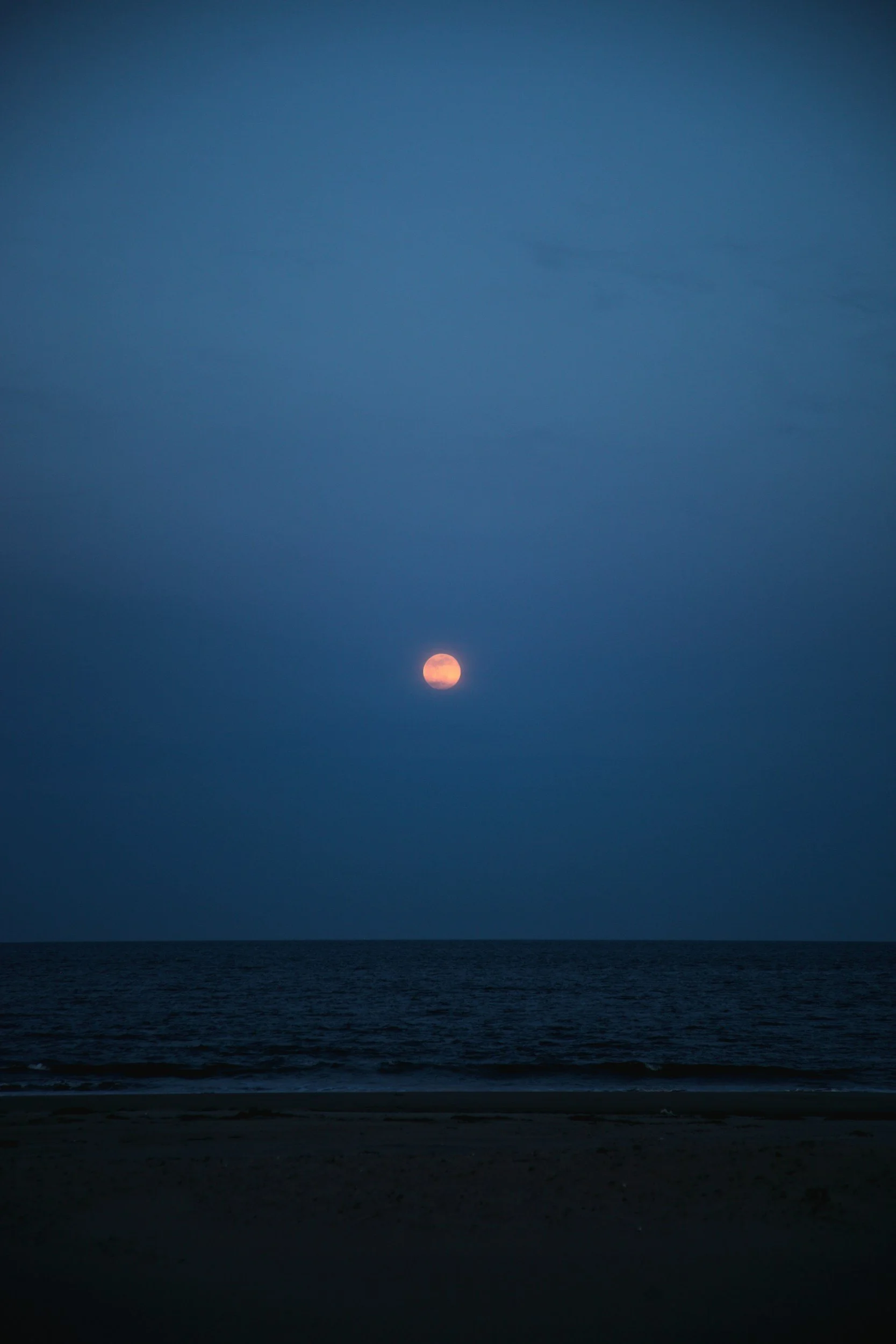 A moon rising over a calm ocean under a dark blue sky.