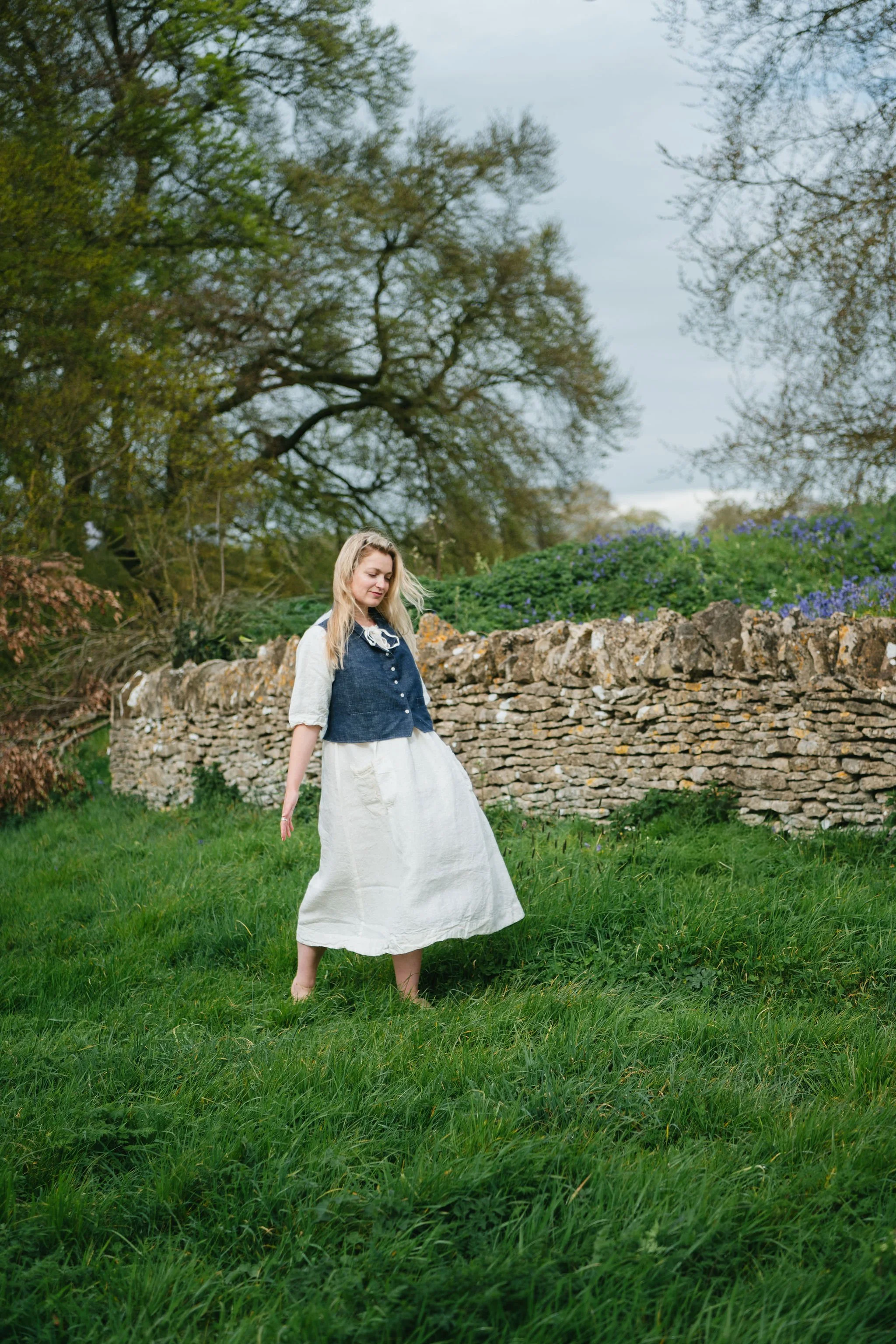 A woman with long blonde hair standing barefoot on a grassy field with a stone wall and trees in the background.