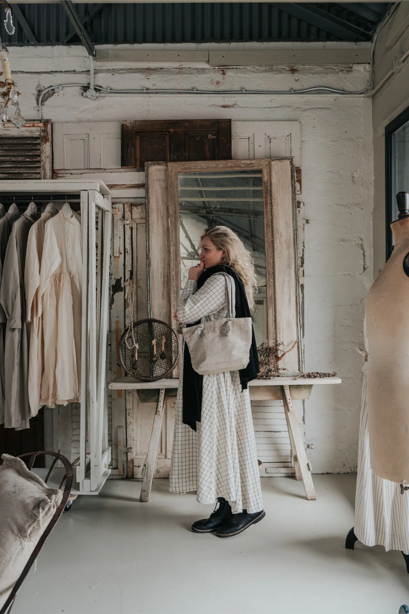 A young woman with blonde hair wearing a long checkered dress, black boots, and a black sweater draped over her shoulders, stands in a vintage clothing store looking at herself in a large mirror. The store has rustic decor with worn wooden frames, white painted brick walls, and hanging clothing. A mannequin and clothing rack are visible, and the woman carries a tote bag.