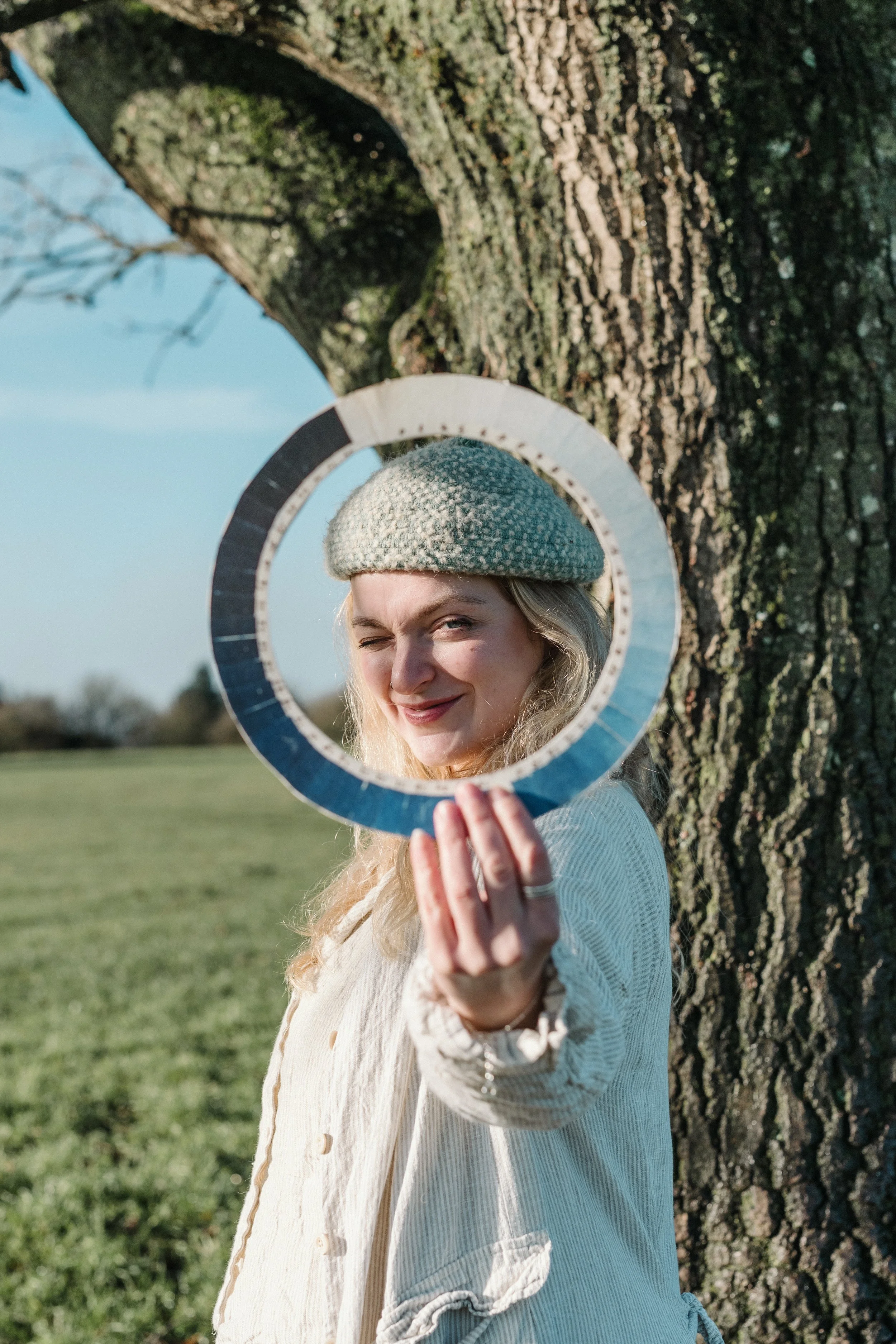 A woman with long blond hair, wearing a beige knitted beret and a light-colored cardigan, is smiling and winking at the camera while holding a round mirror in front of her face. She is standing outdoors beside a large tree with a textured bark, with a grassy field and blue sky in the background.