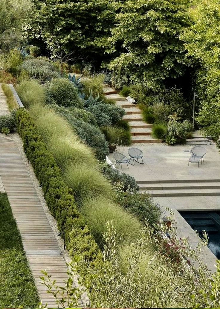 Terraced garden with lush greenery, wooden path, and patio with chairs.