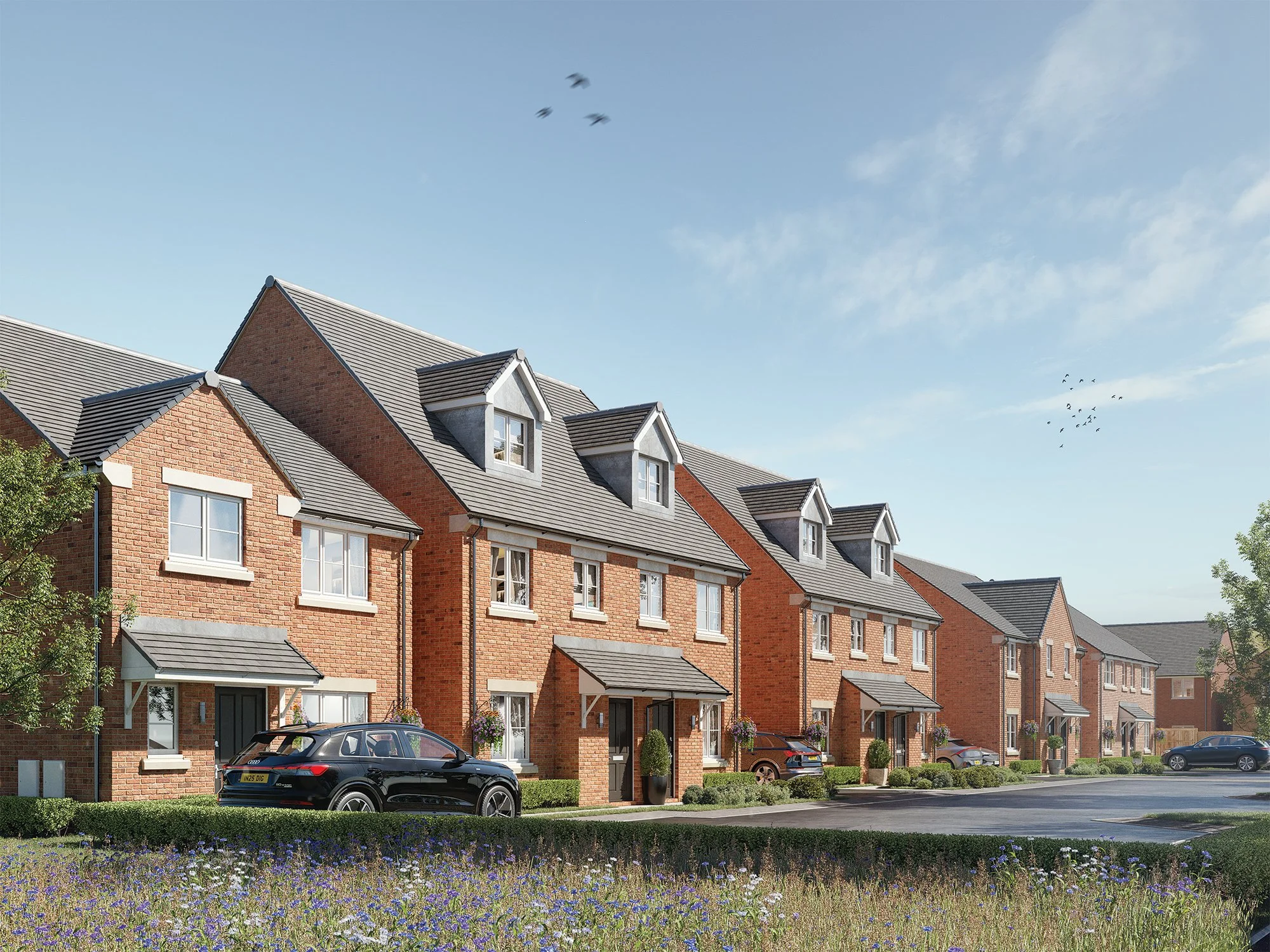 Row of brick townhouses with cars parked in front, front yards with plants, and a grassy area with wildflowers, under a clear blue sky with some clouds and birds flying.
