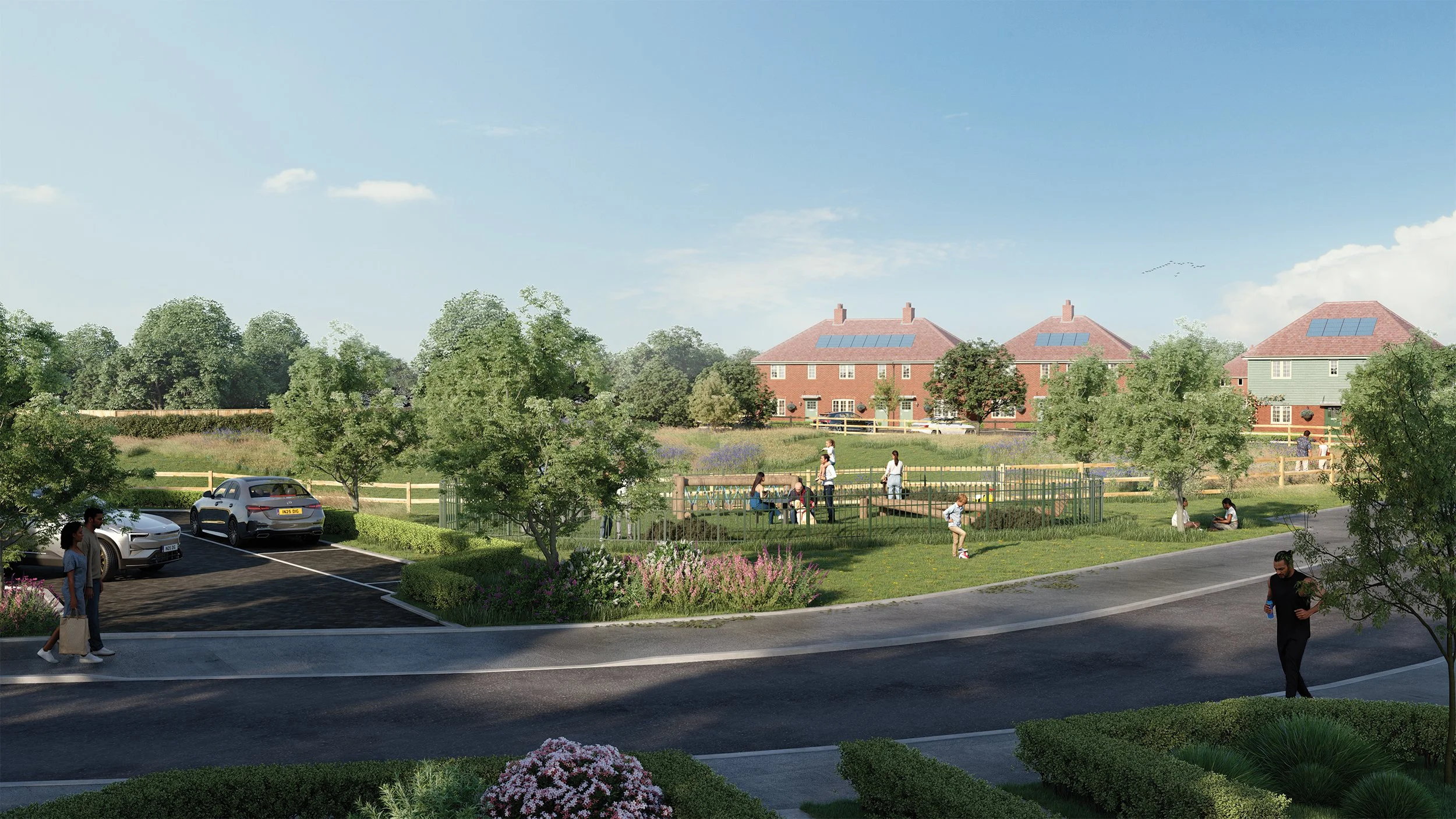 A residential area with a parking lot, walking paths, and houses in the background. Several people are walking, sitting, and playing in the park under a clear blue sky with a few clouds.