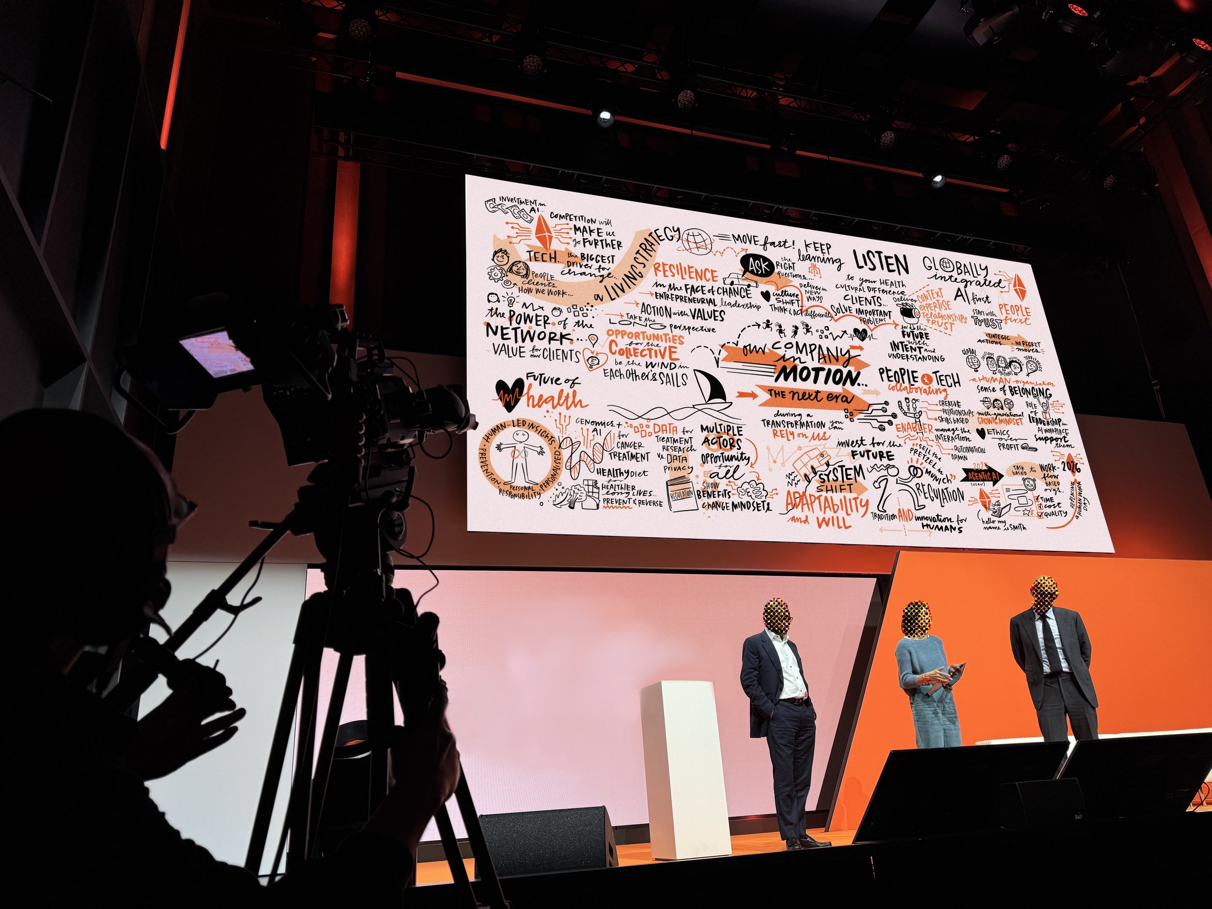 A conference or presentation event with three speakers on stage and a large screen behind them displaying a detailed infographic in black and orange ink.